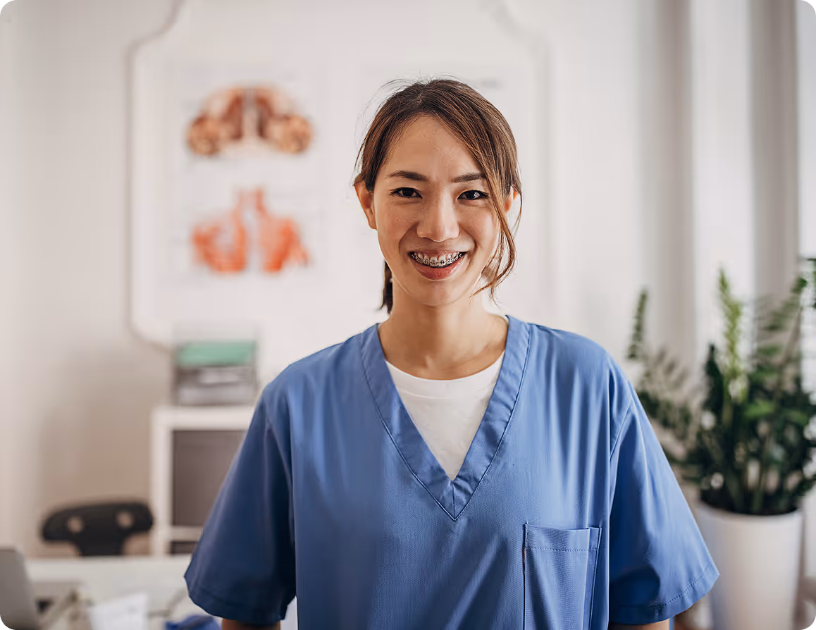 Smiling medical professional wearing braces and blue scrubs in a bright office with plants and anatomy chart.
