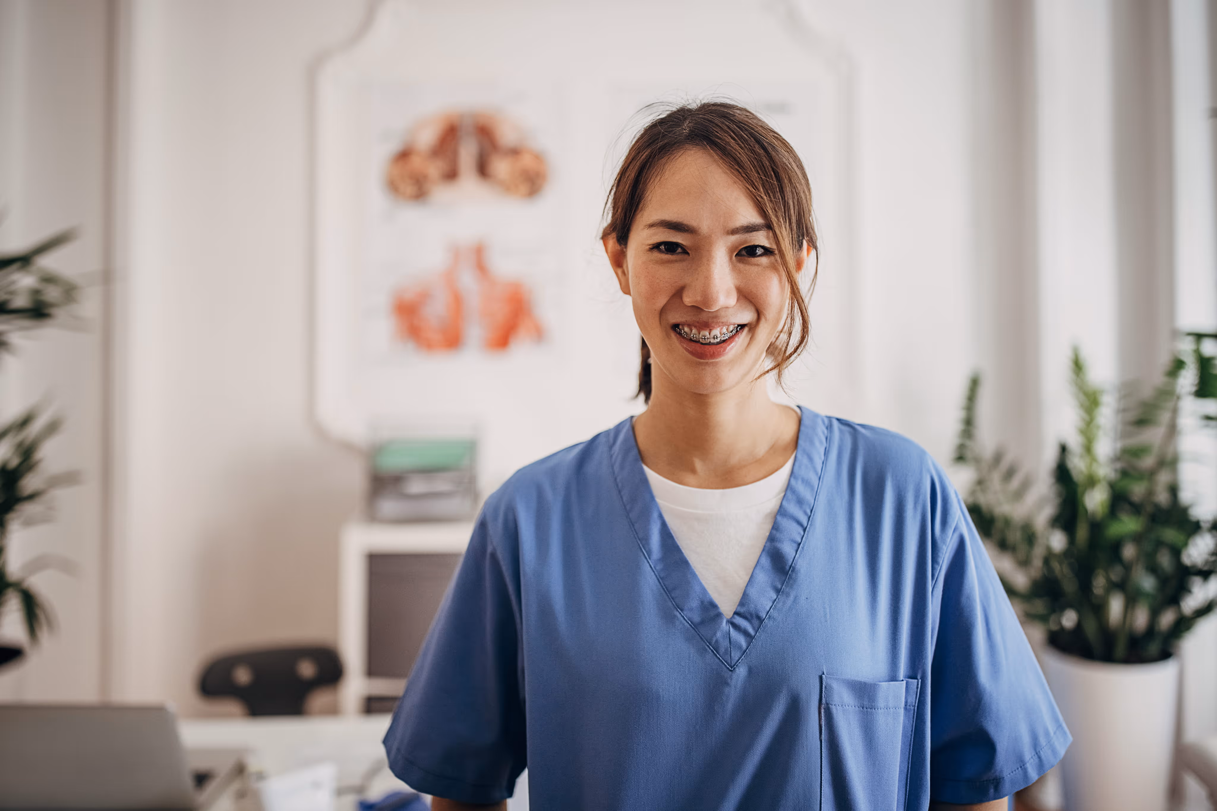 Smiling medical professional wearing braces and blue scrubs in a bright office with plants and anatomy chart.