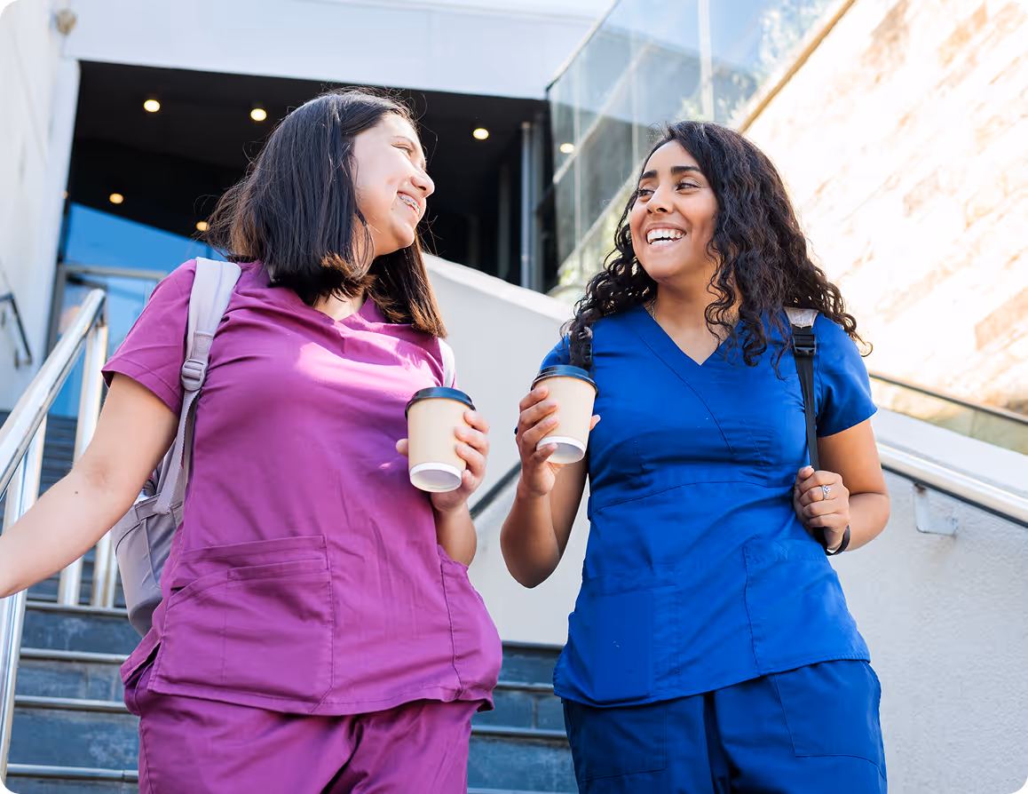 Two female healthcare workers in scrubs walking down stairs holding coffee cups and smiling.