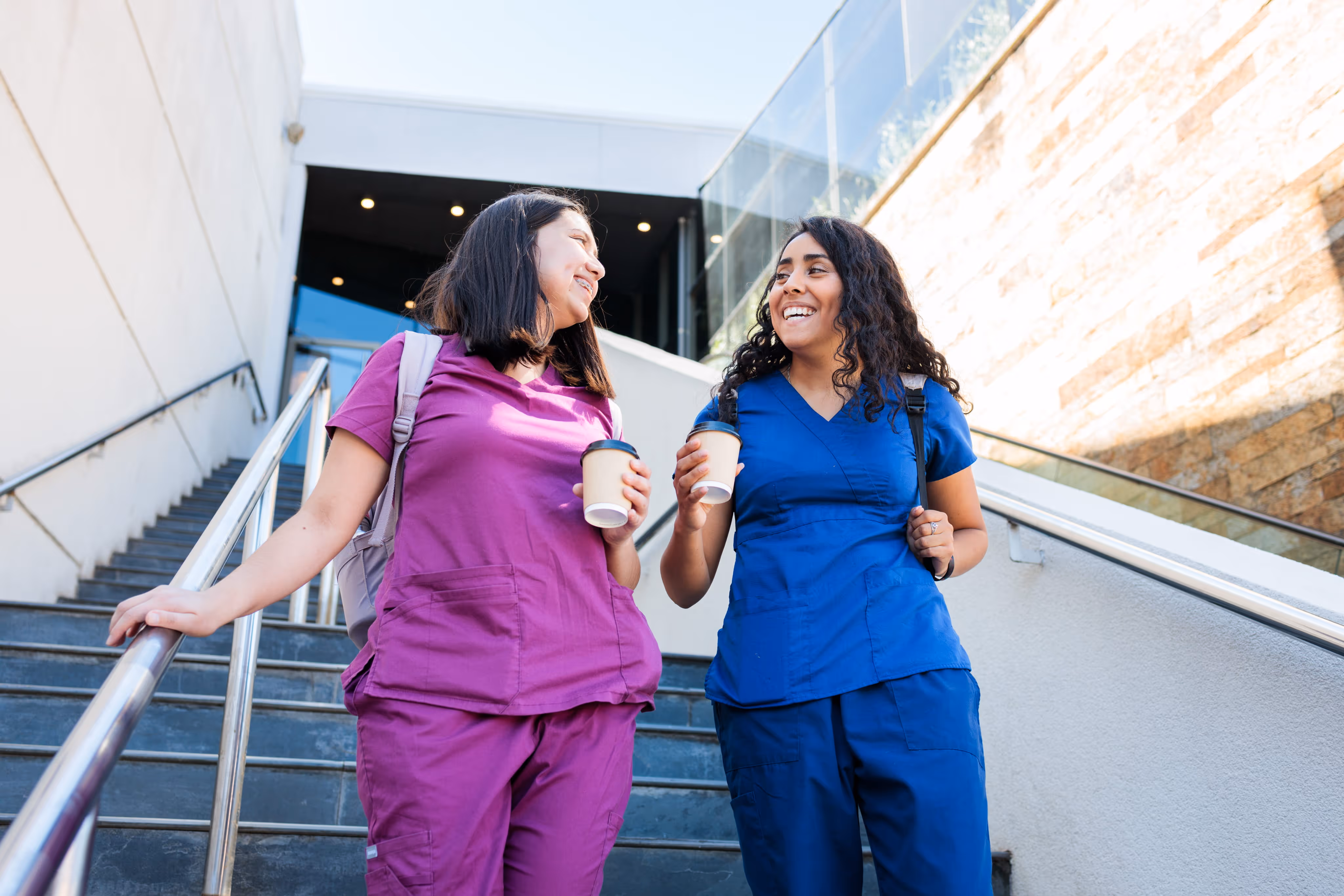 Two female healthcare workers in scrubs walking down stairs holding coffee cups and smiling.