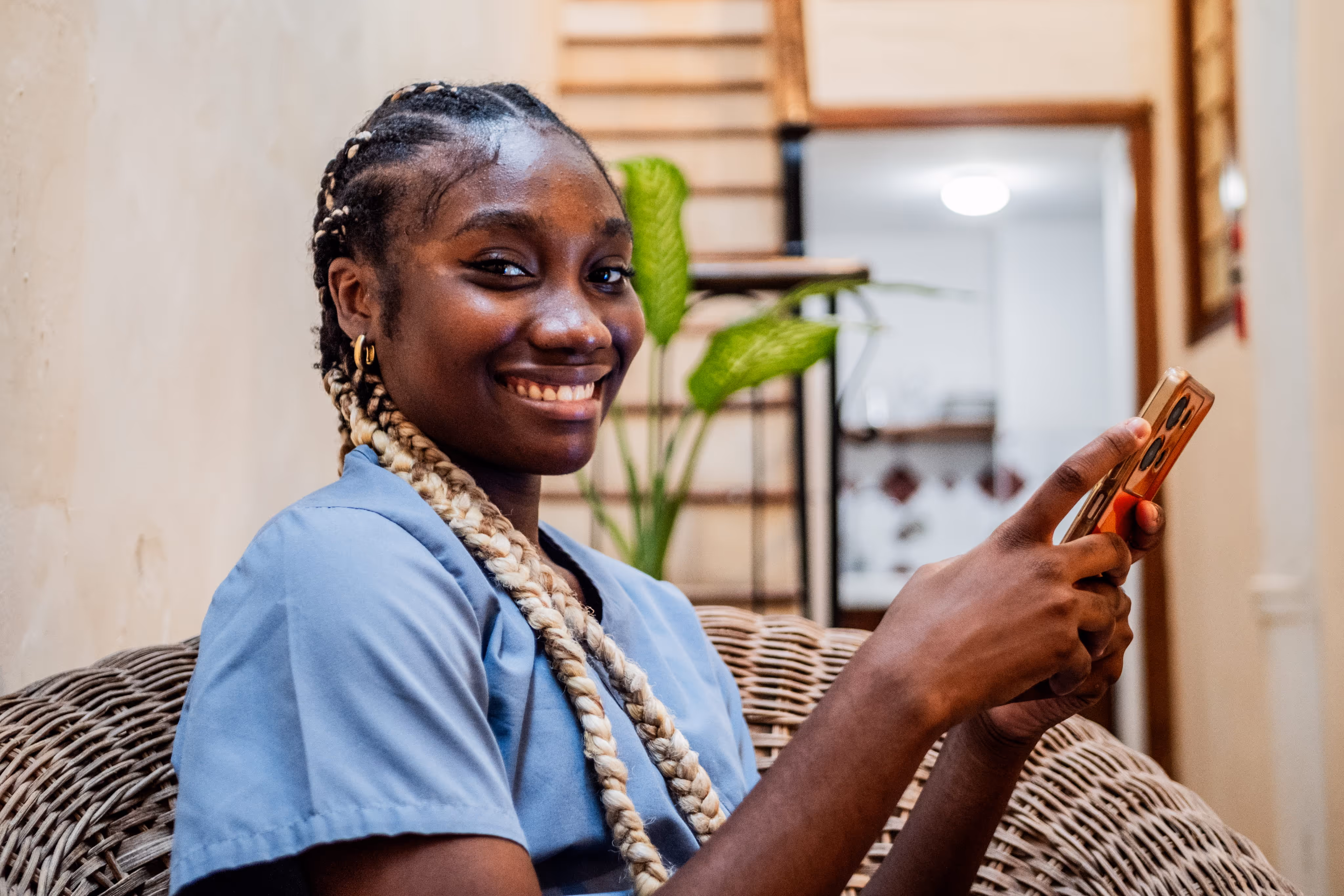 Smiling woman with braided hair sitting on wicker chair holding a smartphone indoors.
