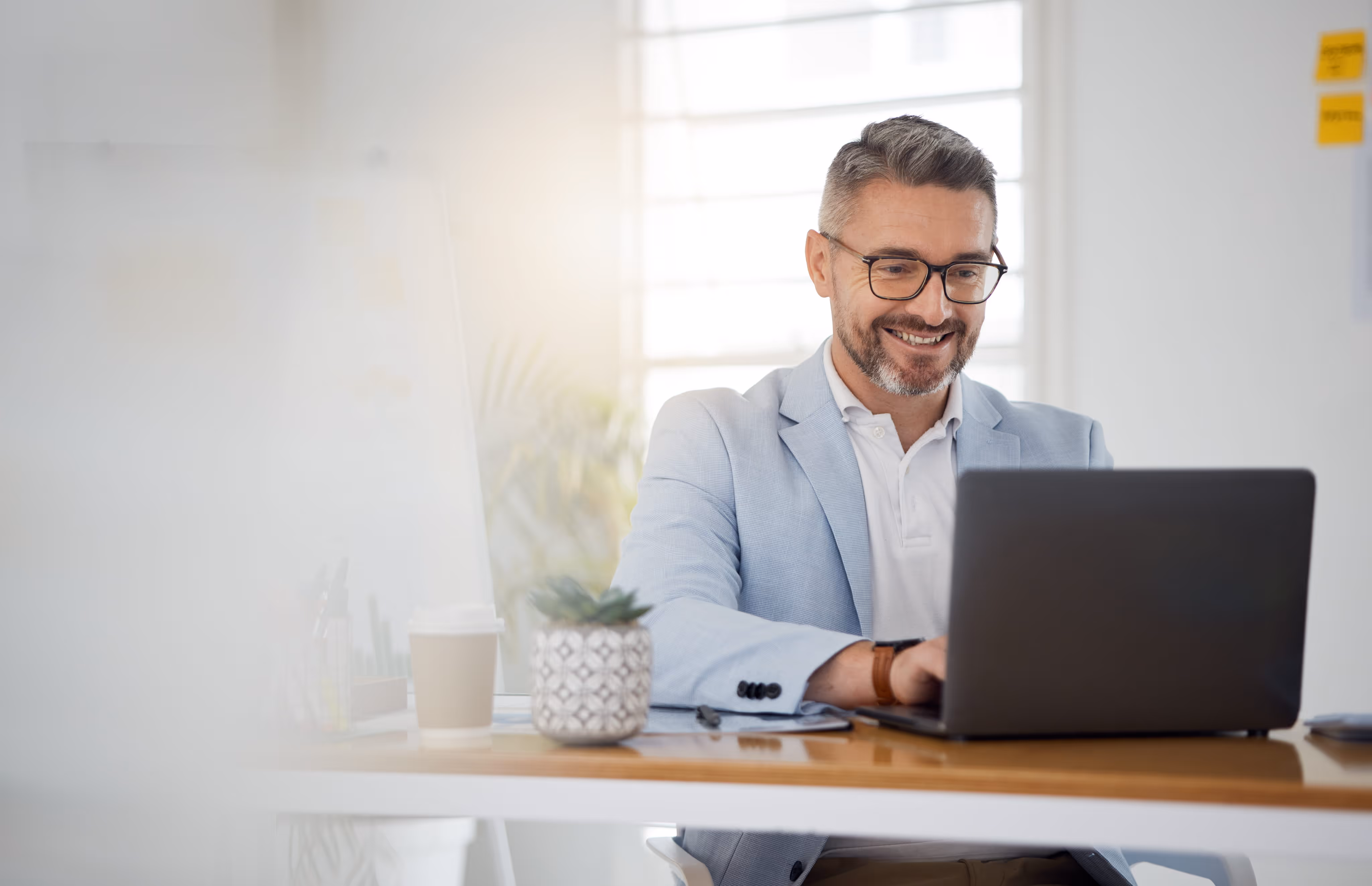 Smiling man in glasses and light blue jacket working on a laptop at a desk with a plant and coffee cup.