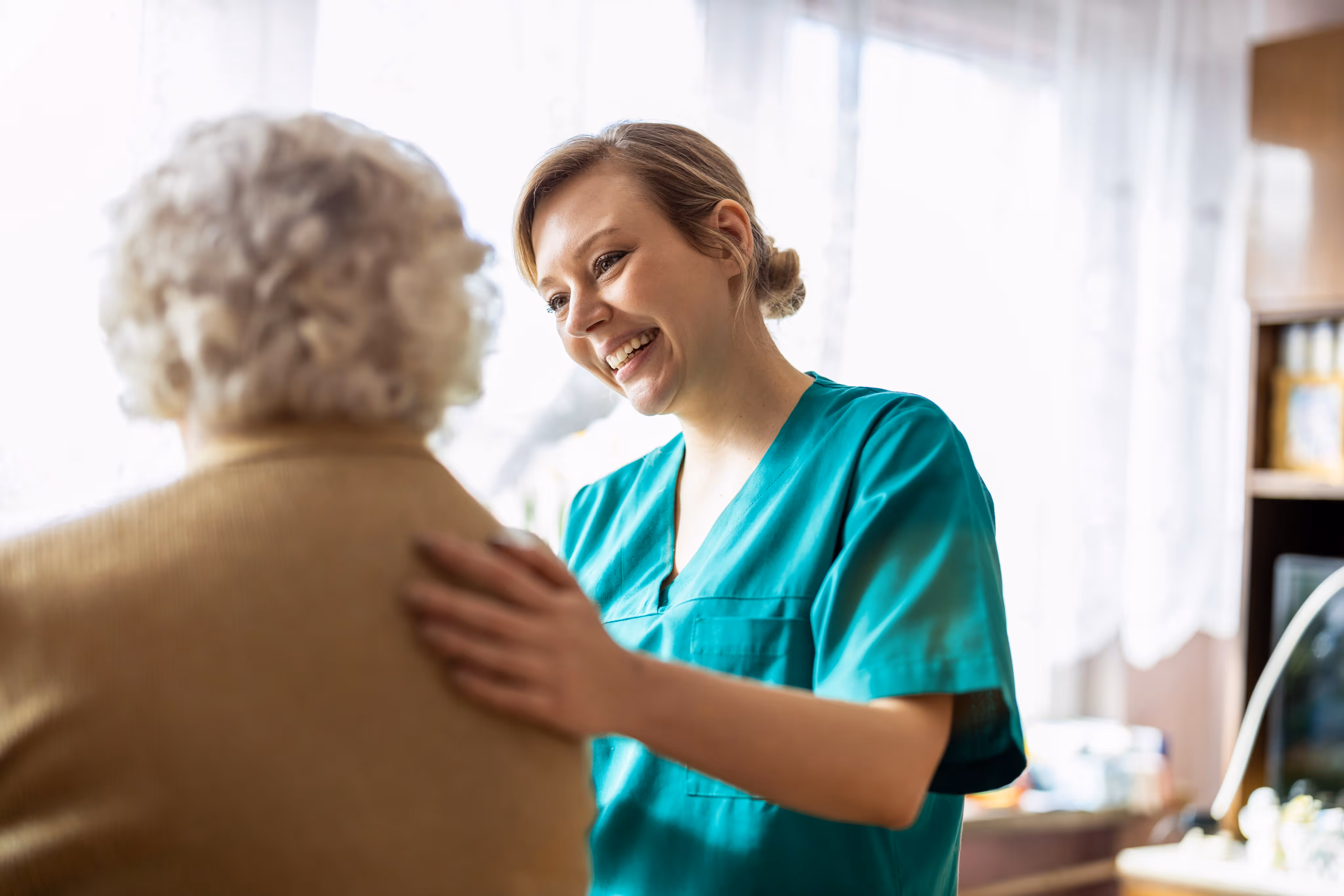 Smiling female nurse in green scrubs comforting elderly person with gray curly hair indoors.