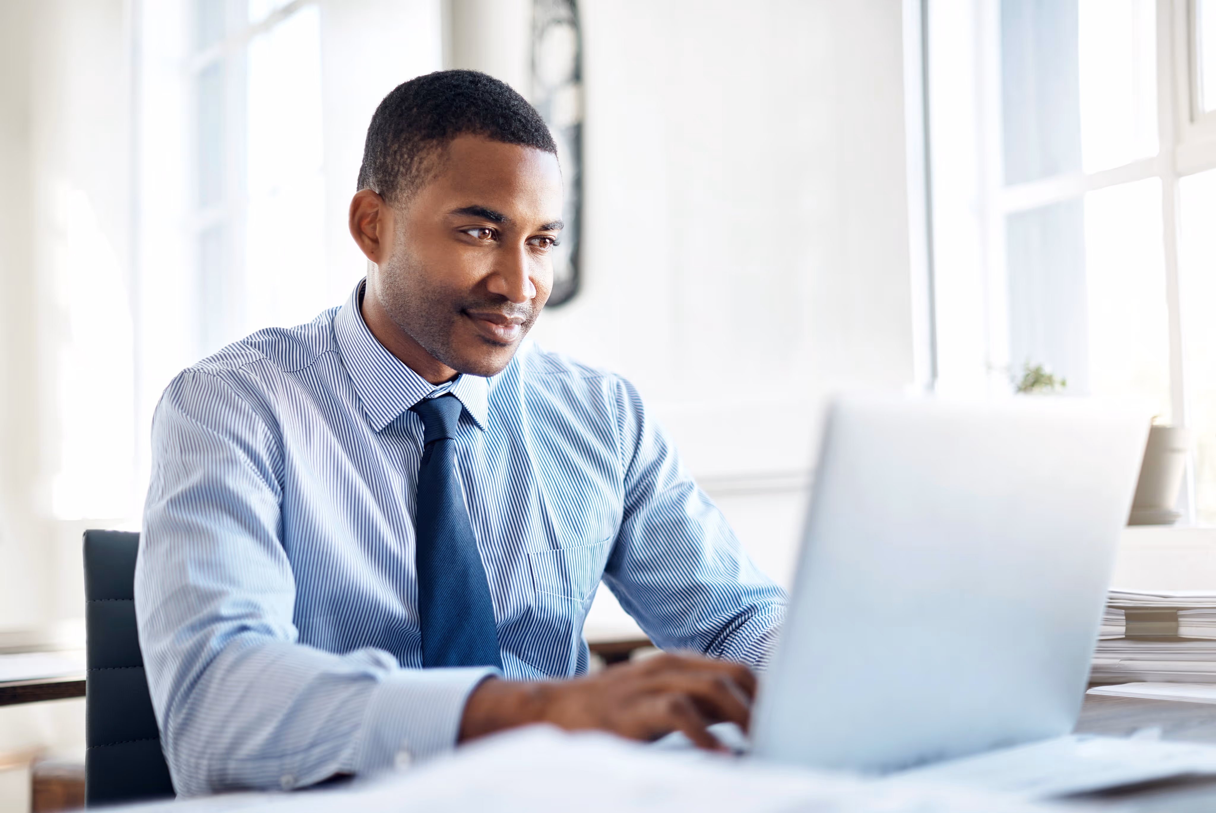 Man in blue striped shirt and tie working on a laptop in a bright office.