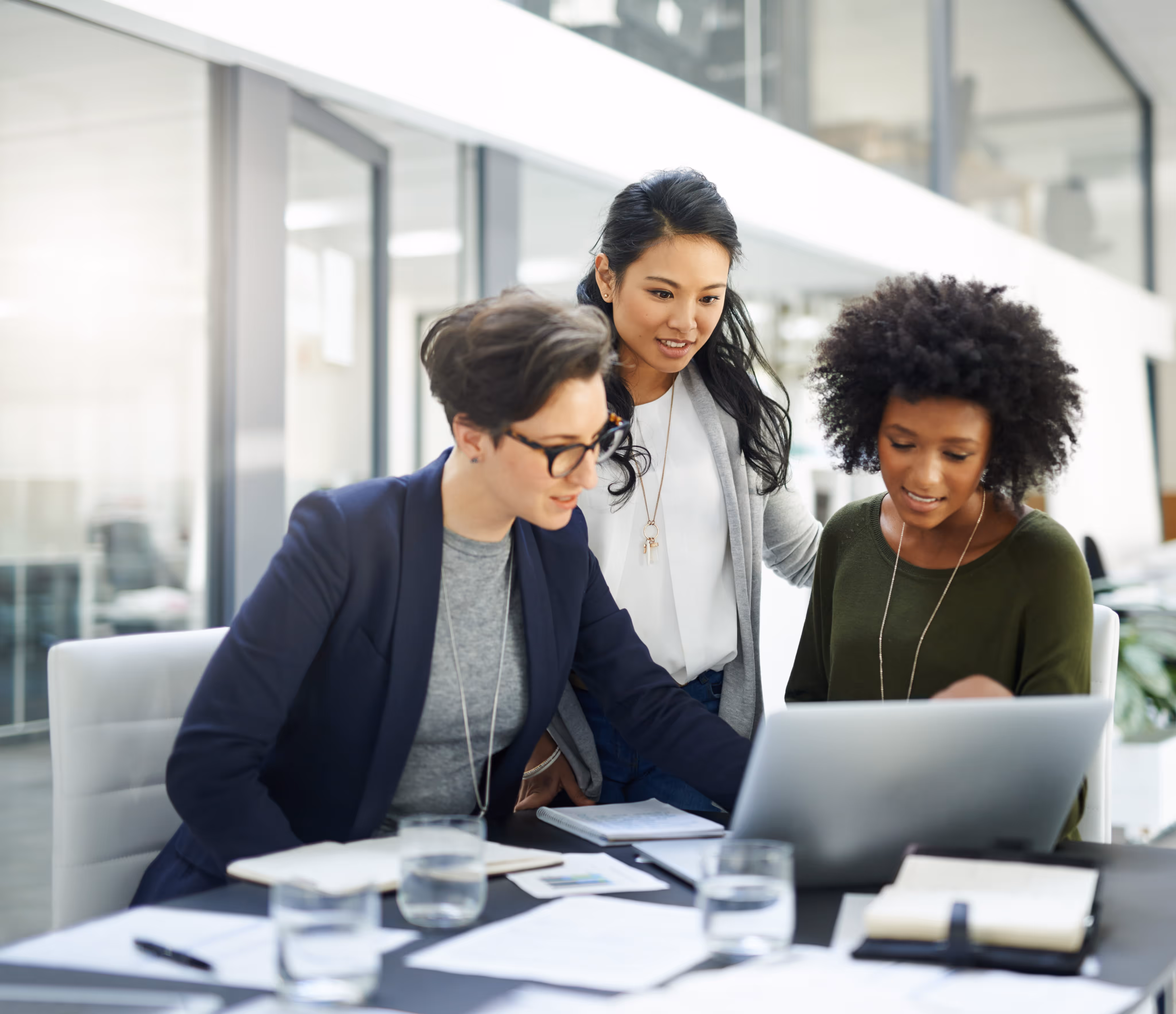 Three diverse women collaborating around a laptop in a modern office workspace.