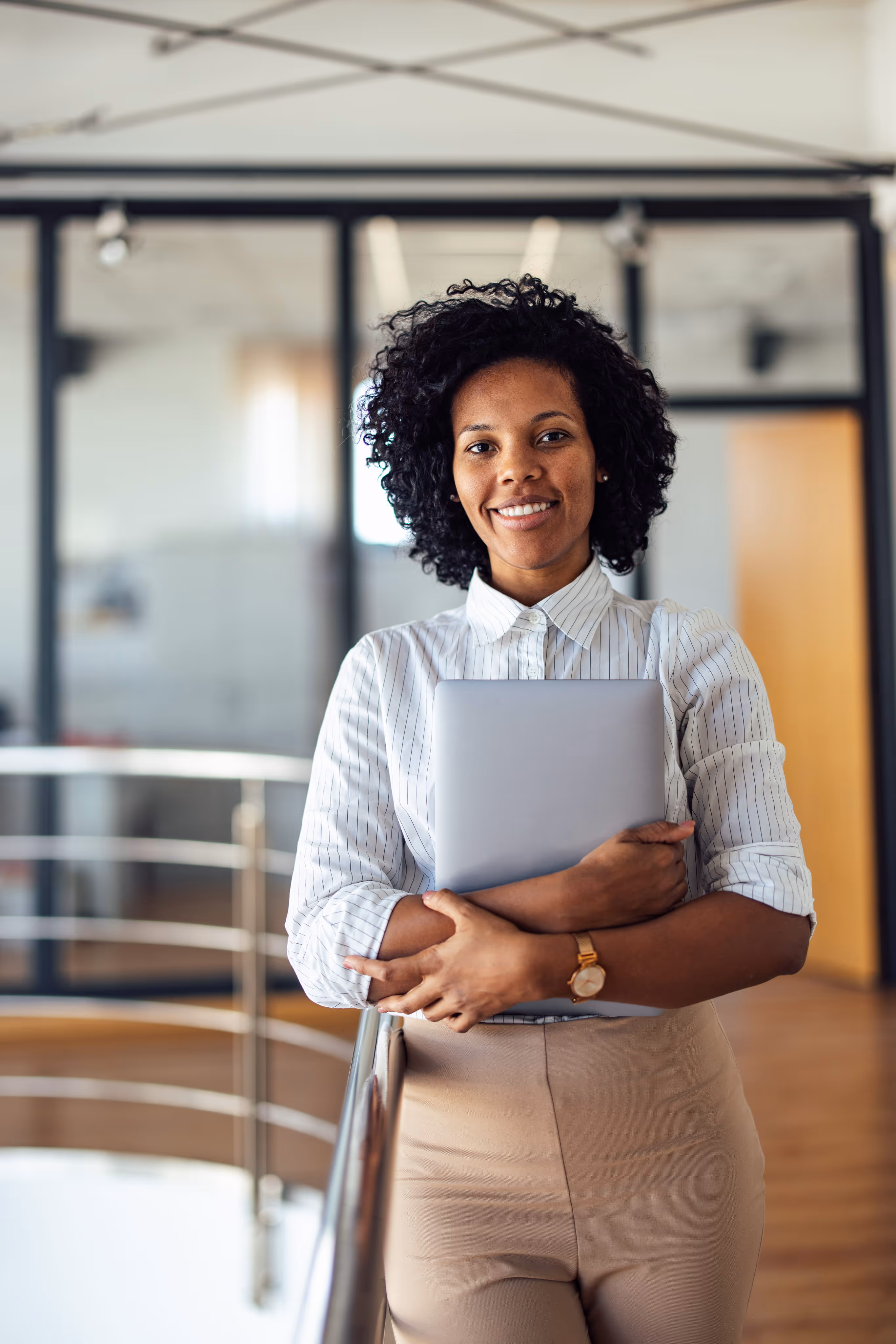 Smiling woman in white shirt holding a closed laptop in a modern office lobby.