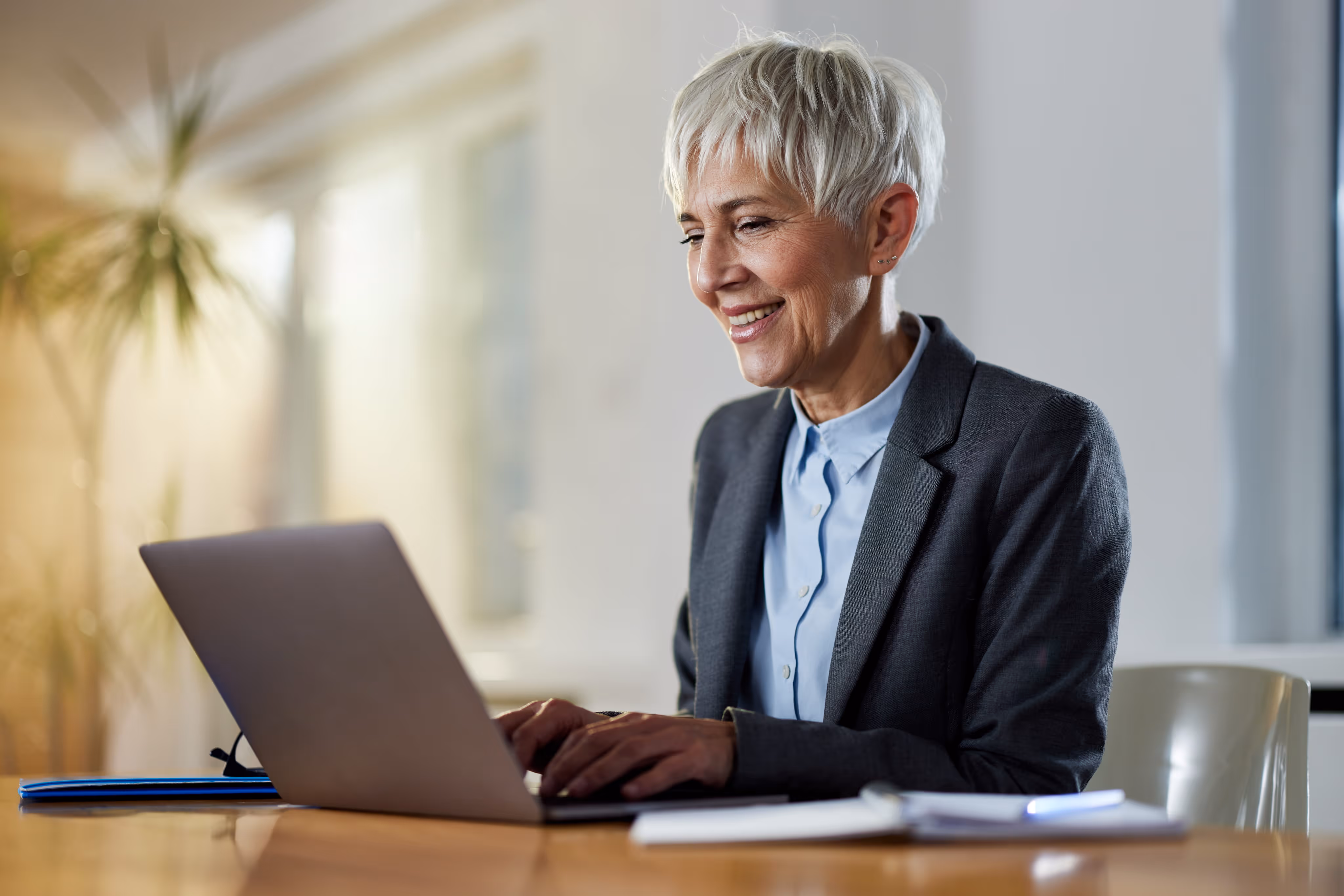 Smiling older woman in business attire working on laptop at wooden desk in bright office.