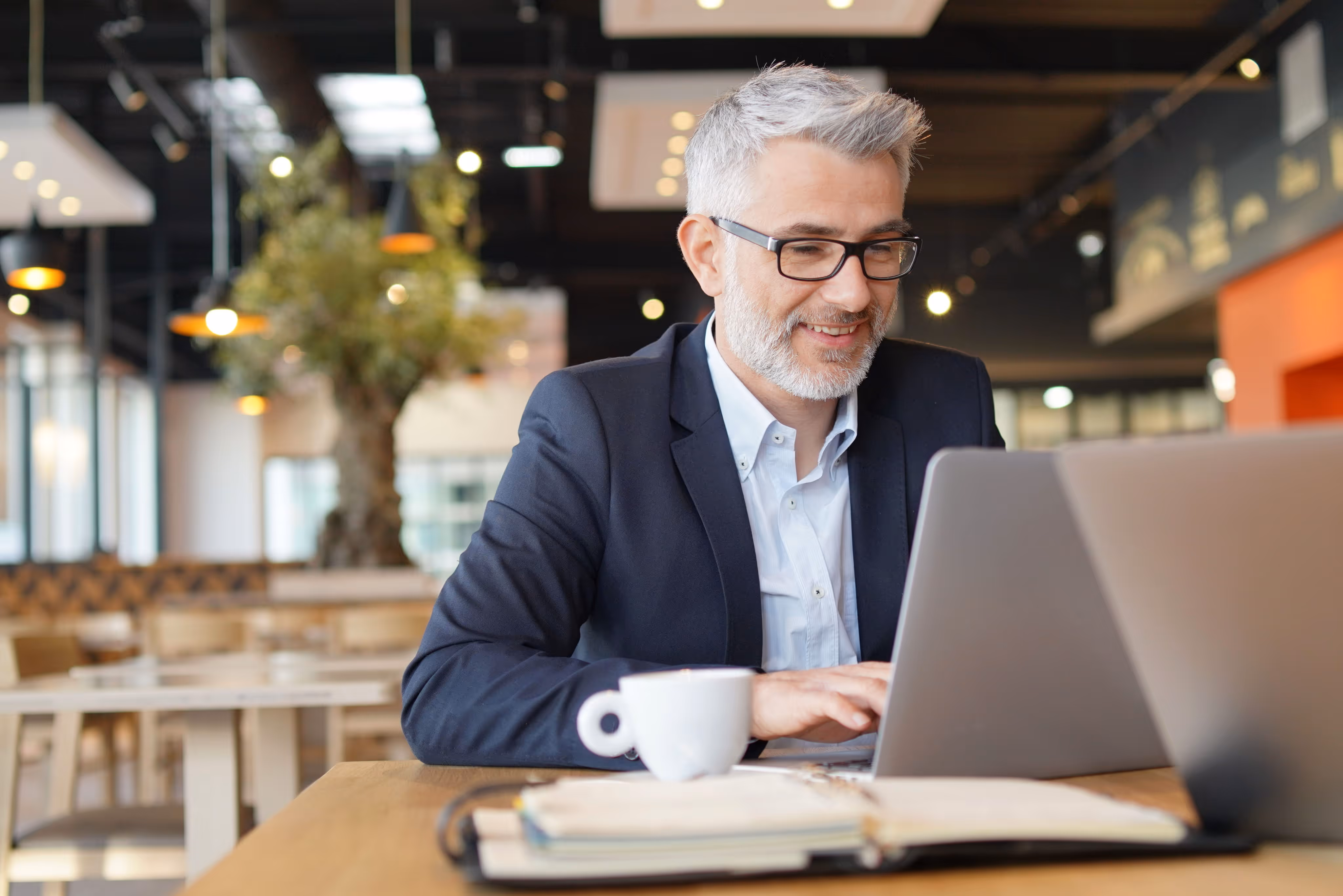 Smiling gray-haired man in glasses and suit typing on laptop at a cafe table with a coffee cup.