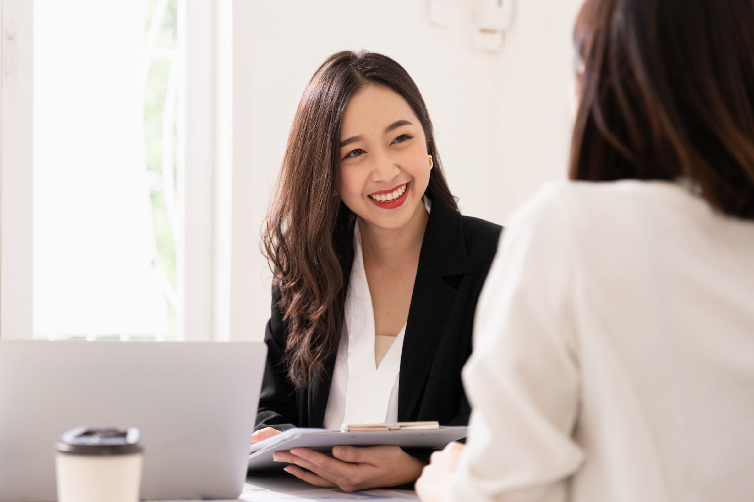 Smiling businesswoman in black blazer holding a clipboard during a meeting with a coworker.