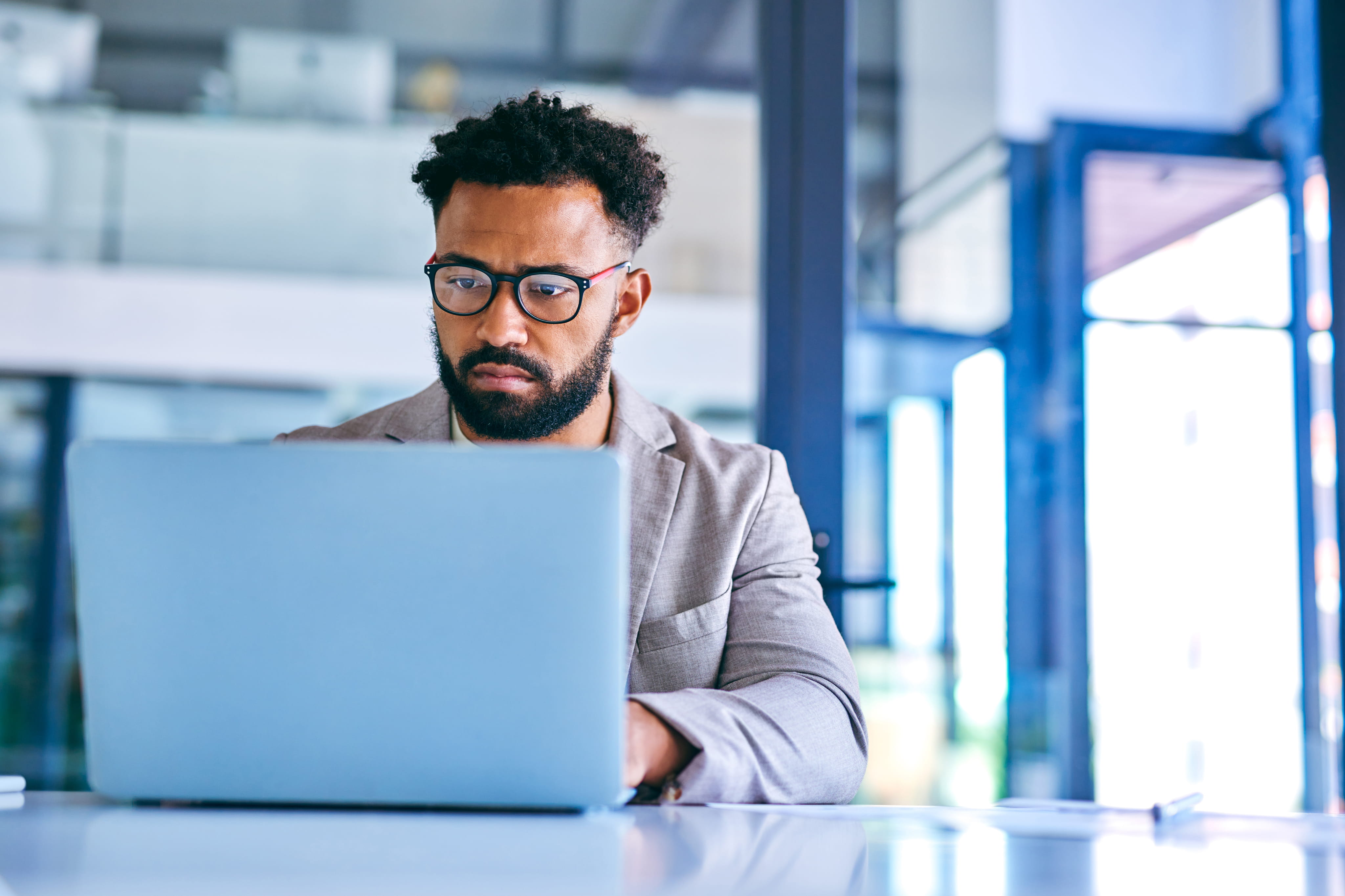 Man with glasses and beard working carefully on a laptop in a modern office.