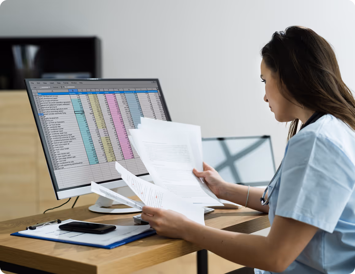 Healthcare professional reviewing documents at desk with spreadsheet data on computer monitor.