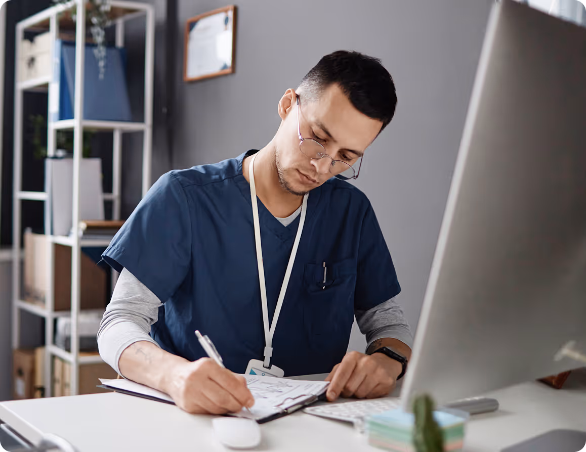 Man in blue scrubs writing on papers at a desk with a large computer monitor.