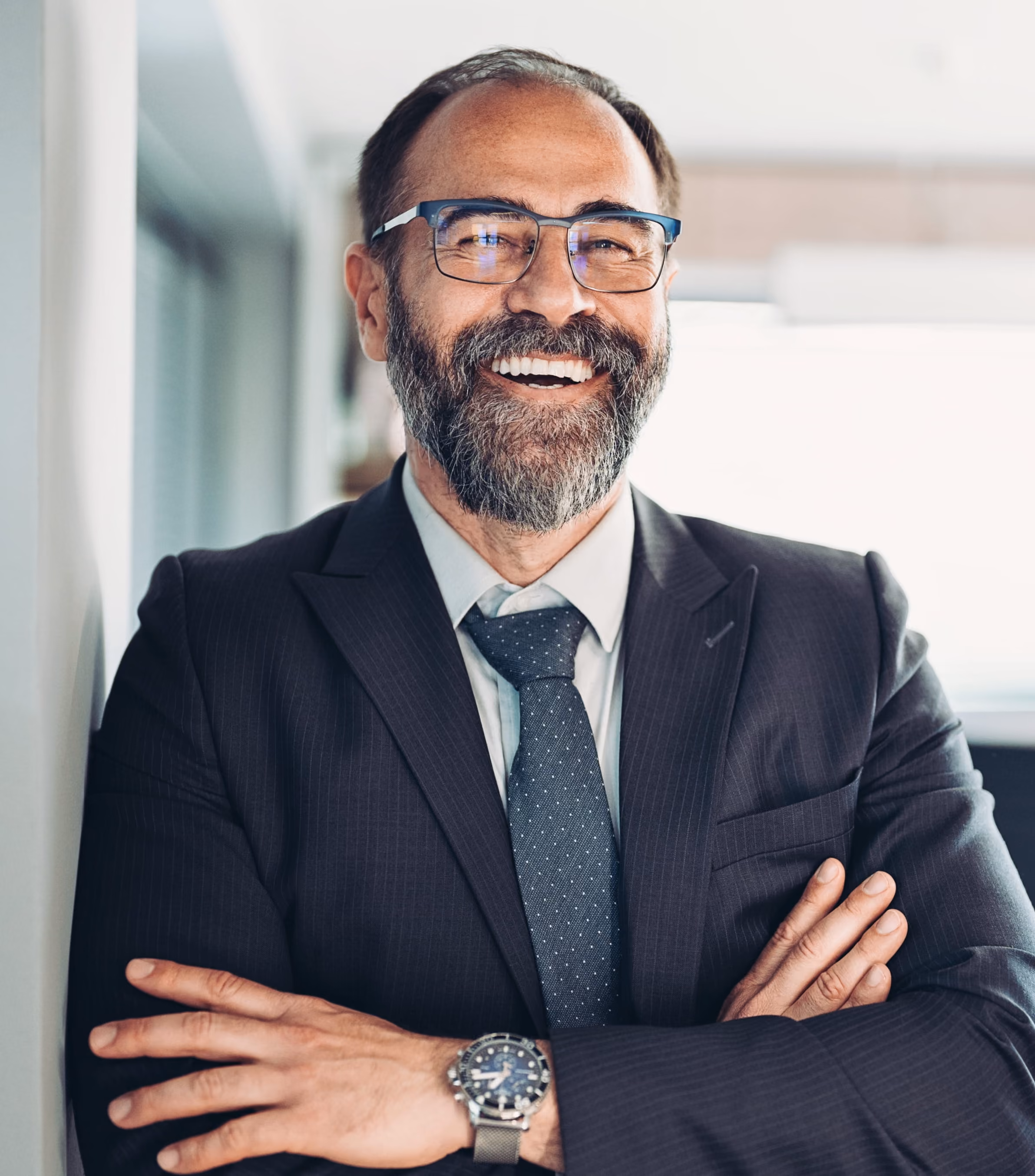 Smiling bearded man in suit and glasses with arms crossed in a modern office setting.