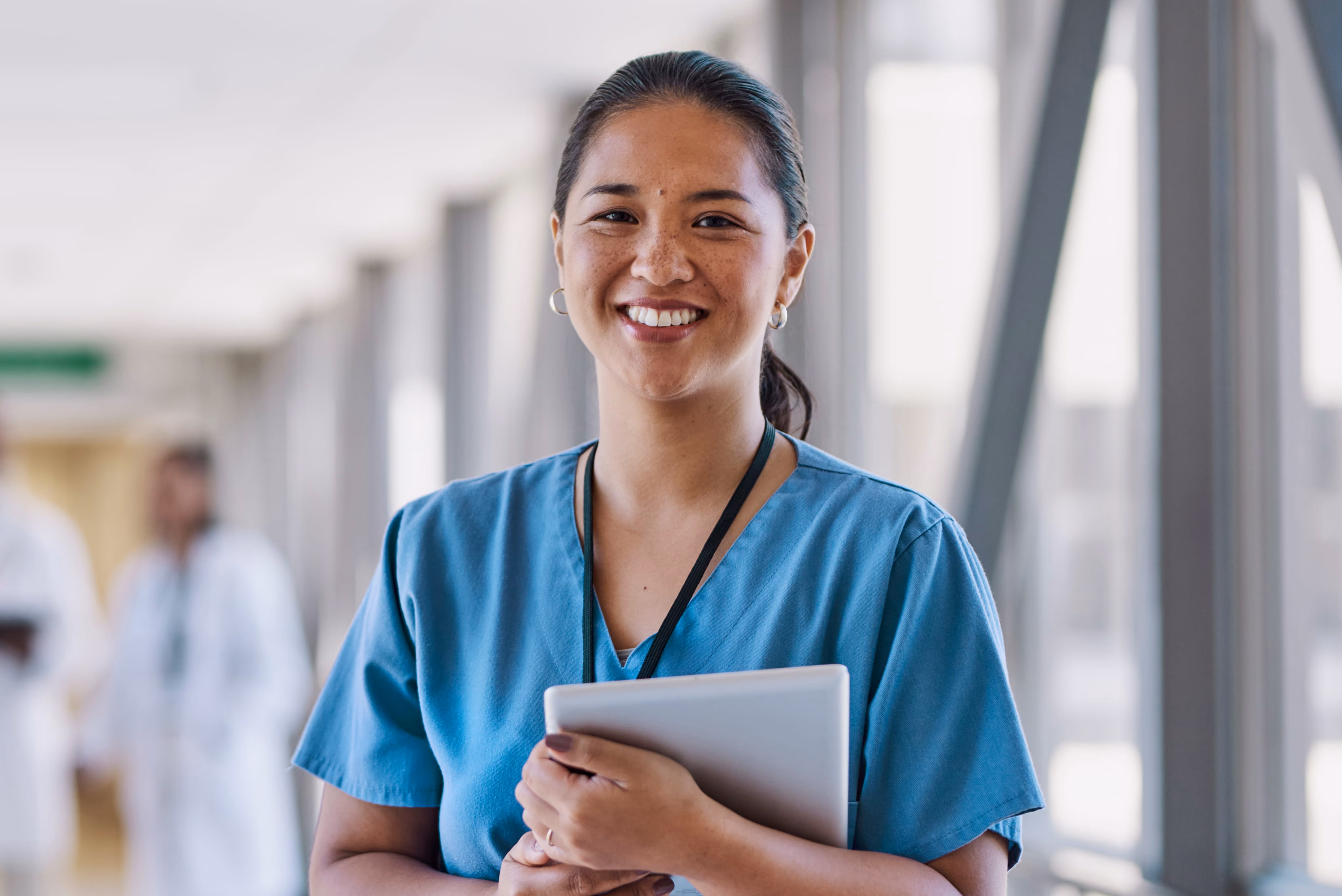 Smiling female nurse in blue scrubs holding a tablet in a hospital corridor.
