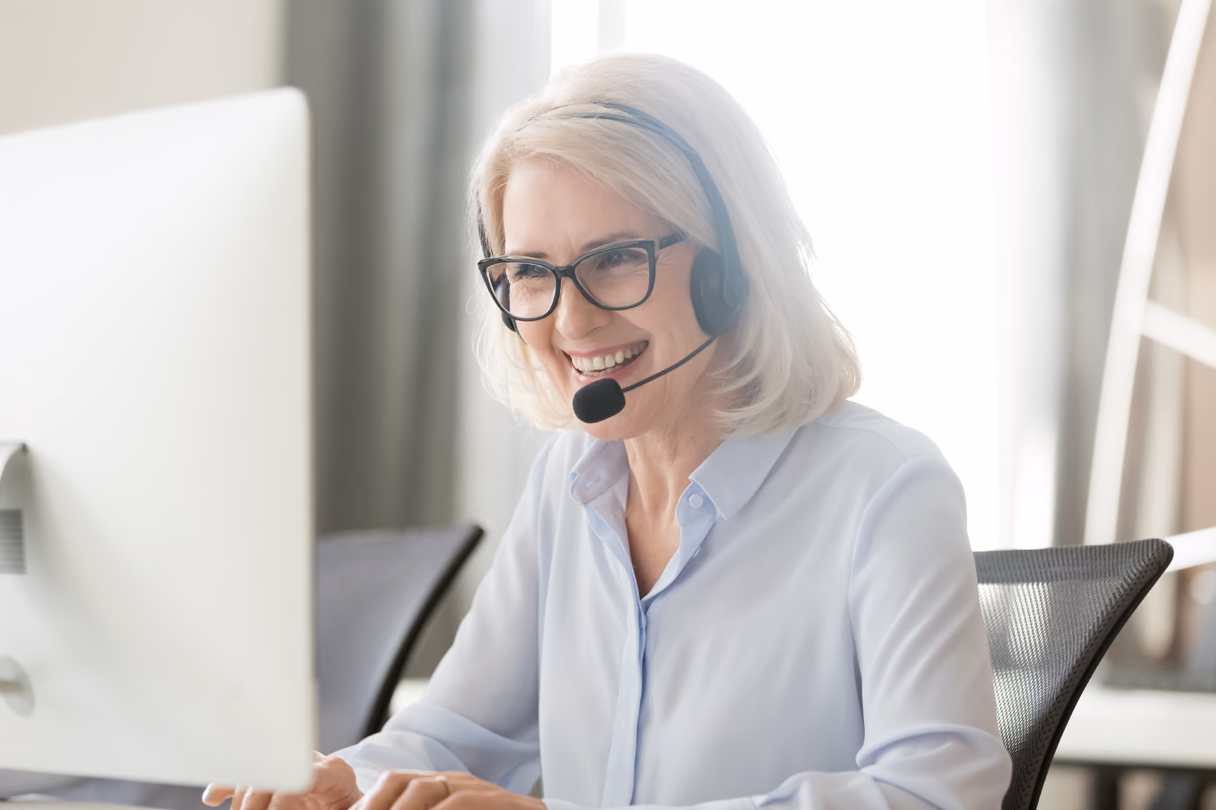 Smiling woman with glasses wearing a headset, working at a computer in an office.