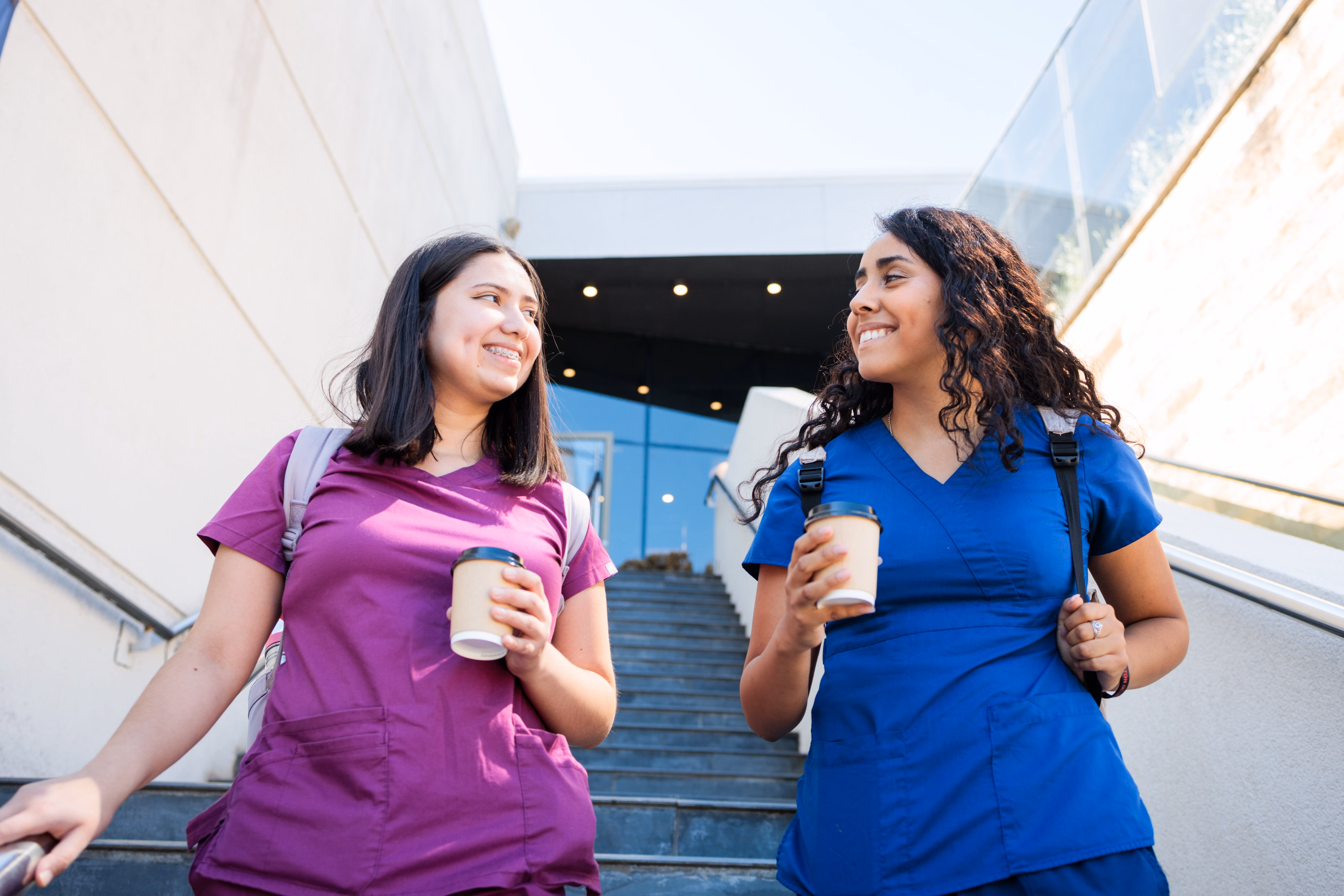 Two smiling women in scrubs holding coffee cups, walking down outdoor stairs, carrying backpacks.