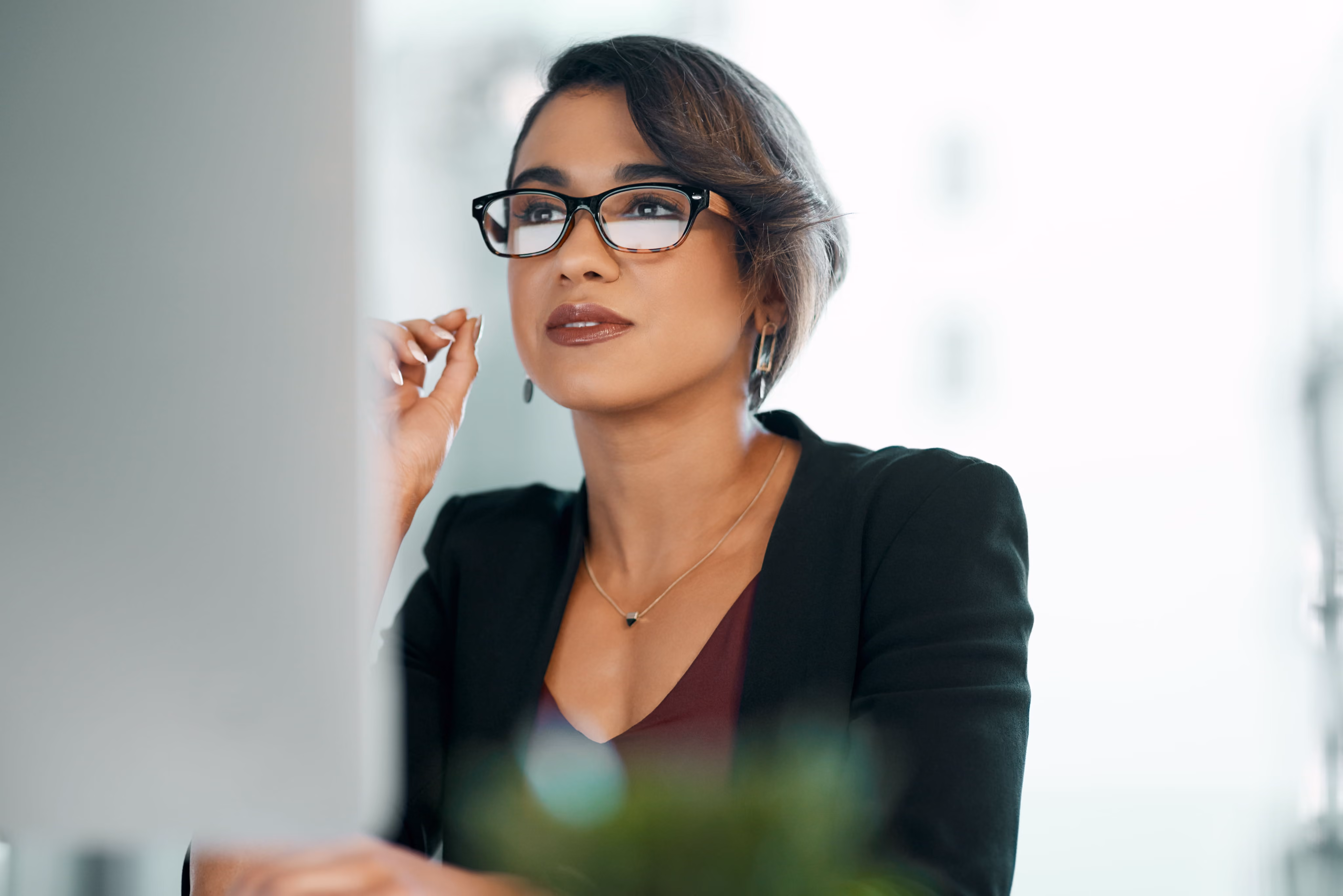 Woman with glasses and short hair looking thoughtfully at a computer screen in an office.