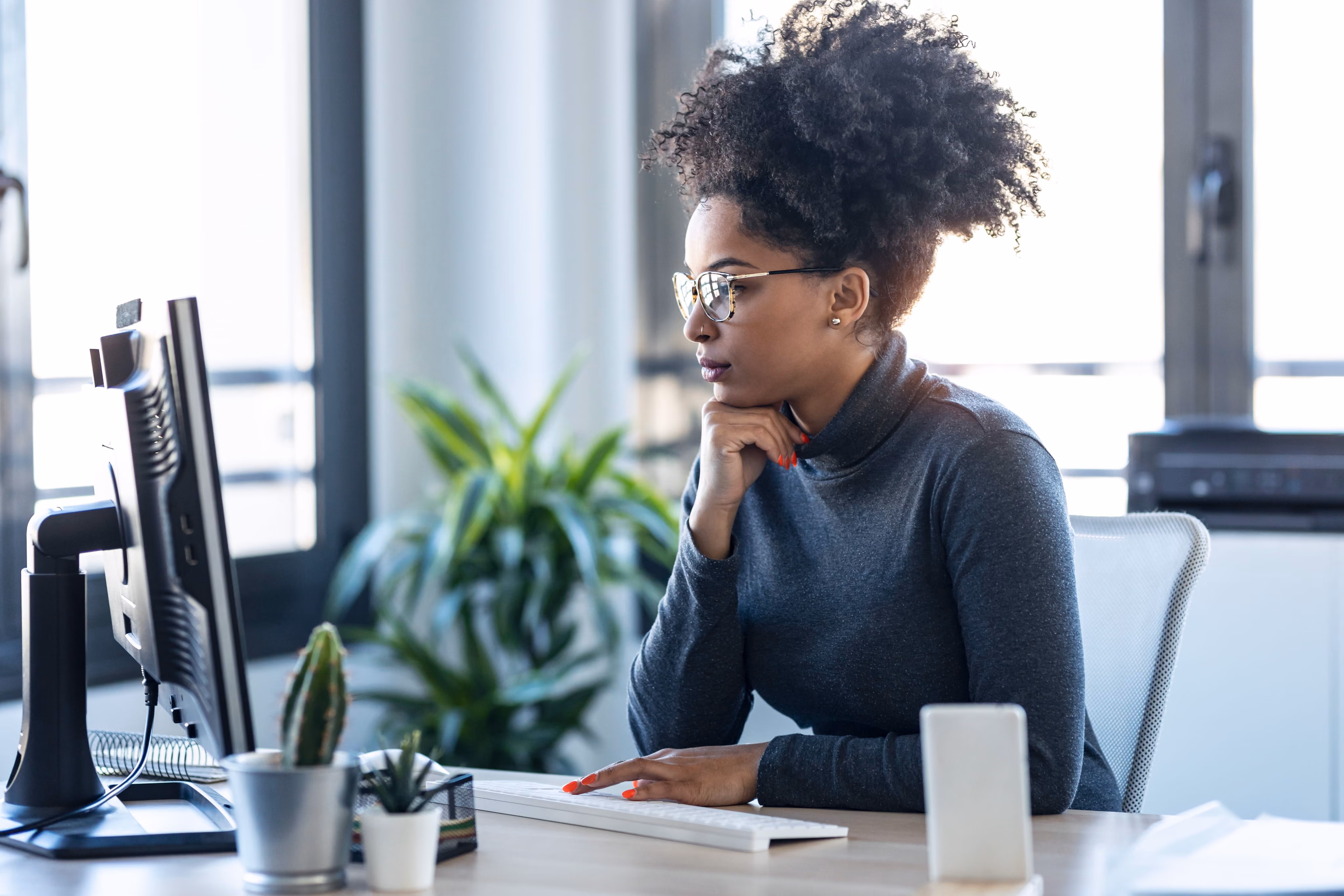 Woman with curly hair and glasses working at a desktop computer in a bright office.