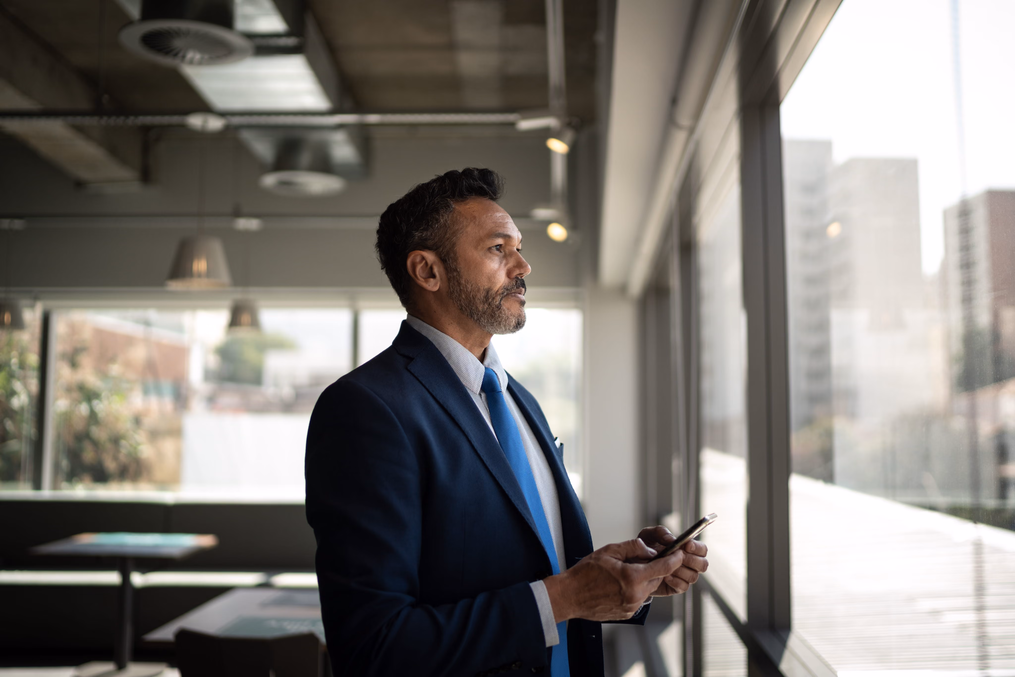 Businessman in suit and blue tie looking out office window while holding a smartphone.