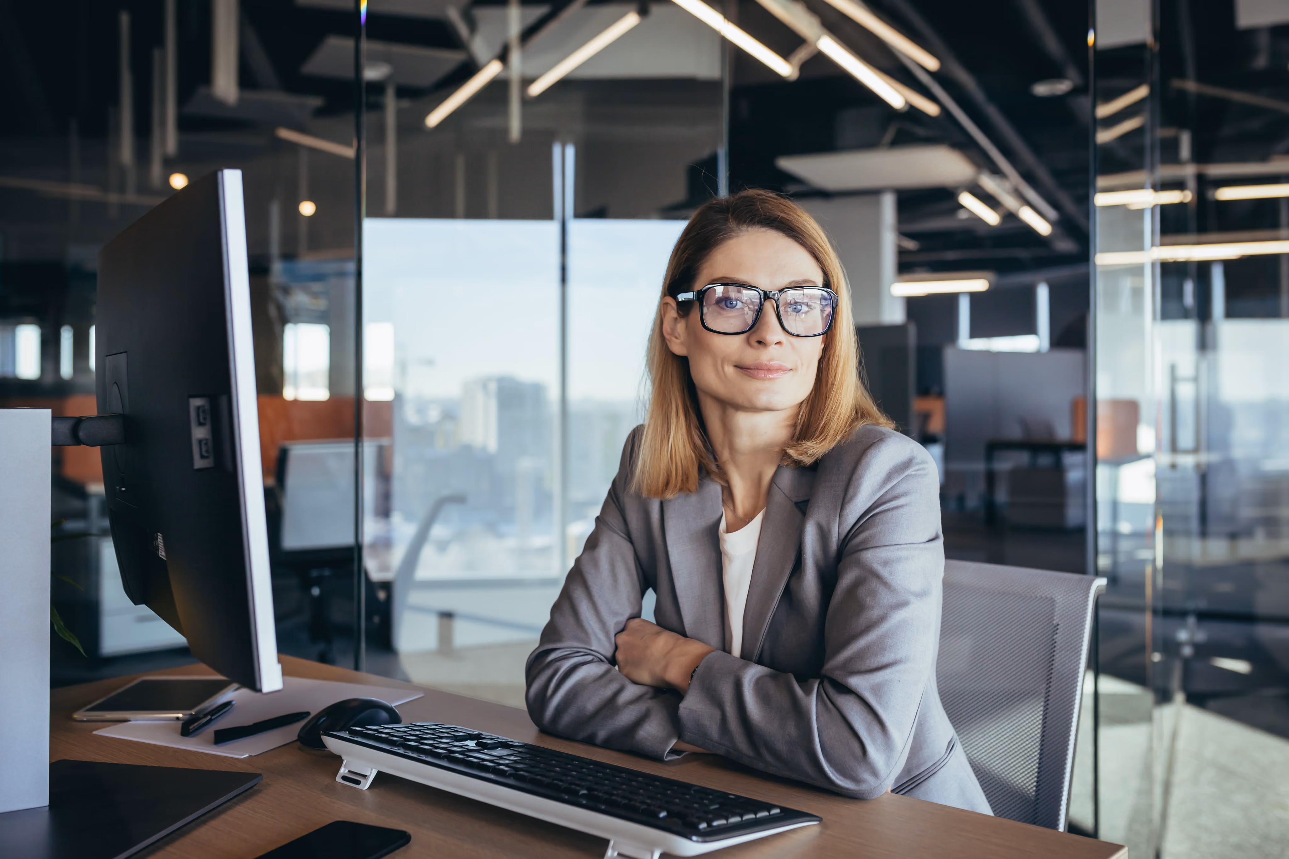 Woman in glasses and gray blazer sitting with crossed arms at a desk with computer in office.