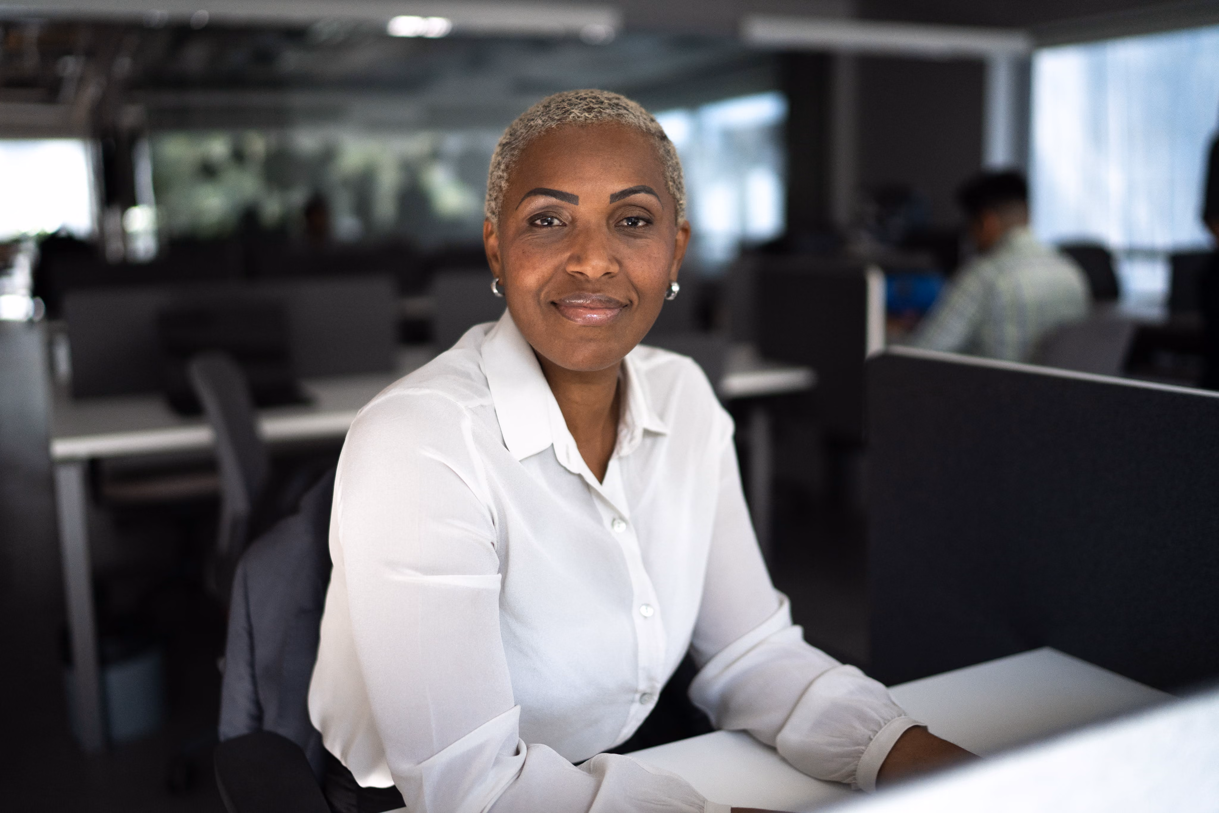 Smiling woman with short blonde hair wearing a white shirt, sitting at a desk in an office.
