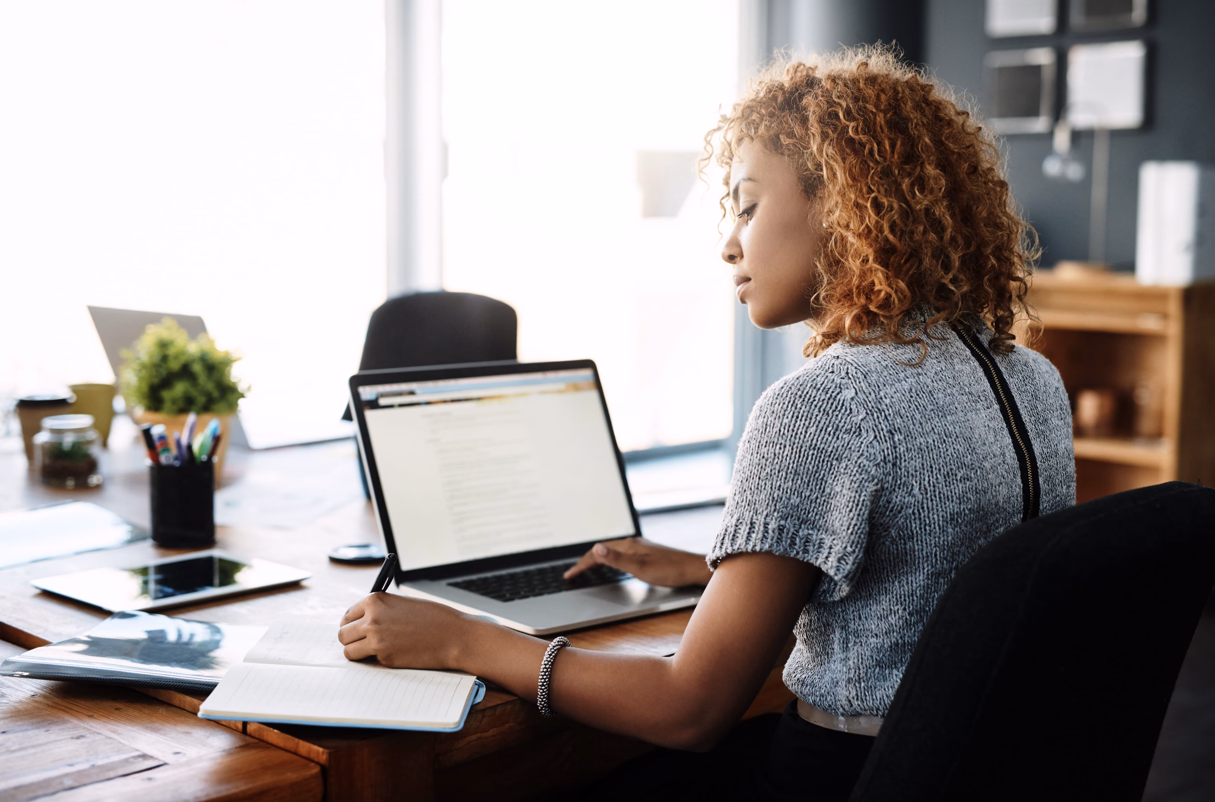 Woman with curly hair sits at a desk writing in a notebook while using a laptop in a bright office.