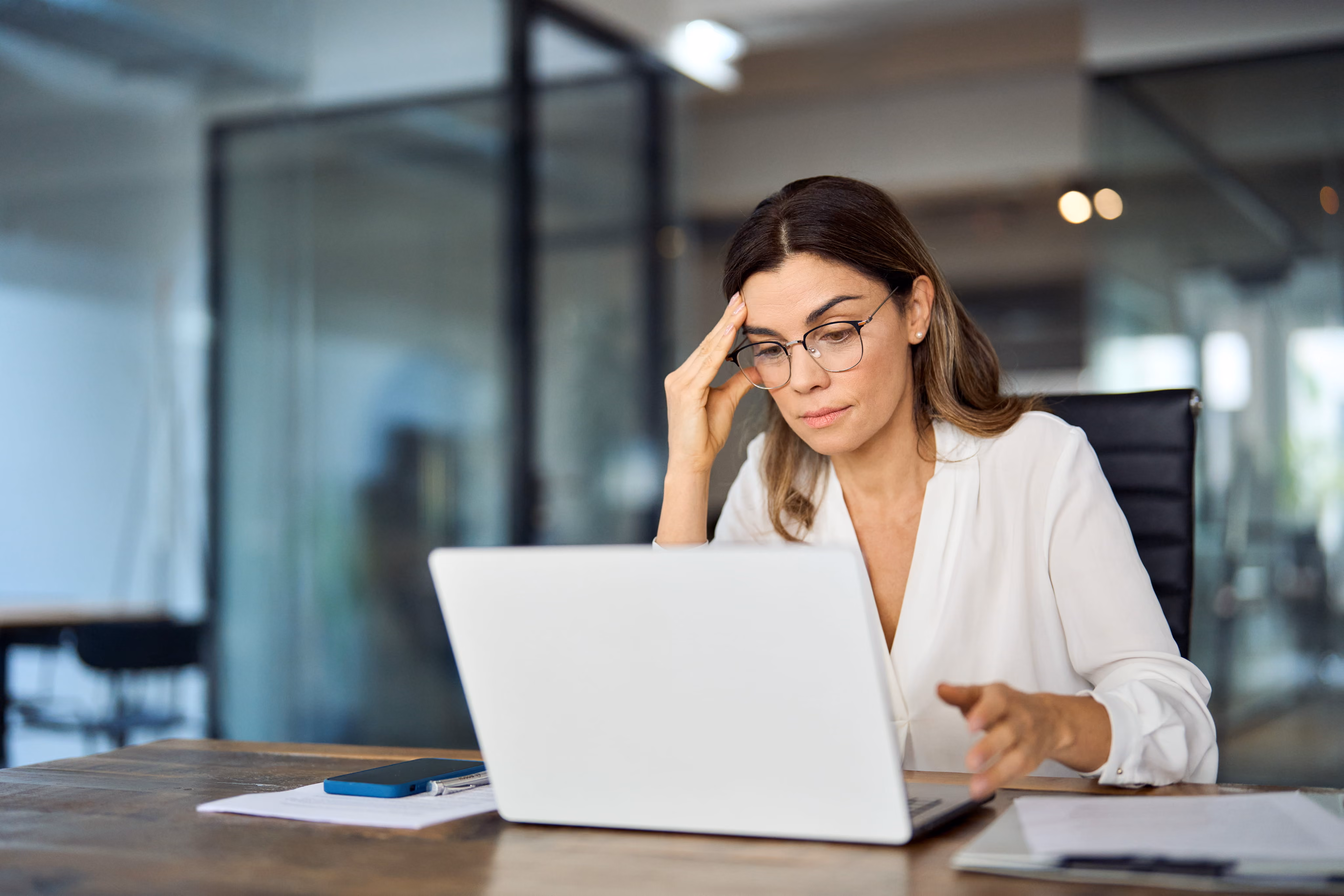 Woman with glasses looking concerned while working on a laptop at a desk with papers and phone.
