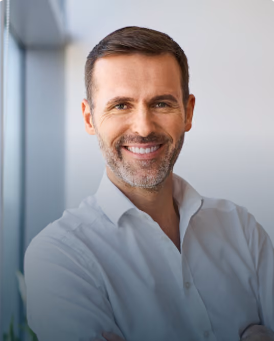 Smiling man with short hair and trimmed beard wearing a white shirt standing by a window.