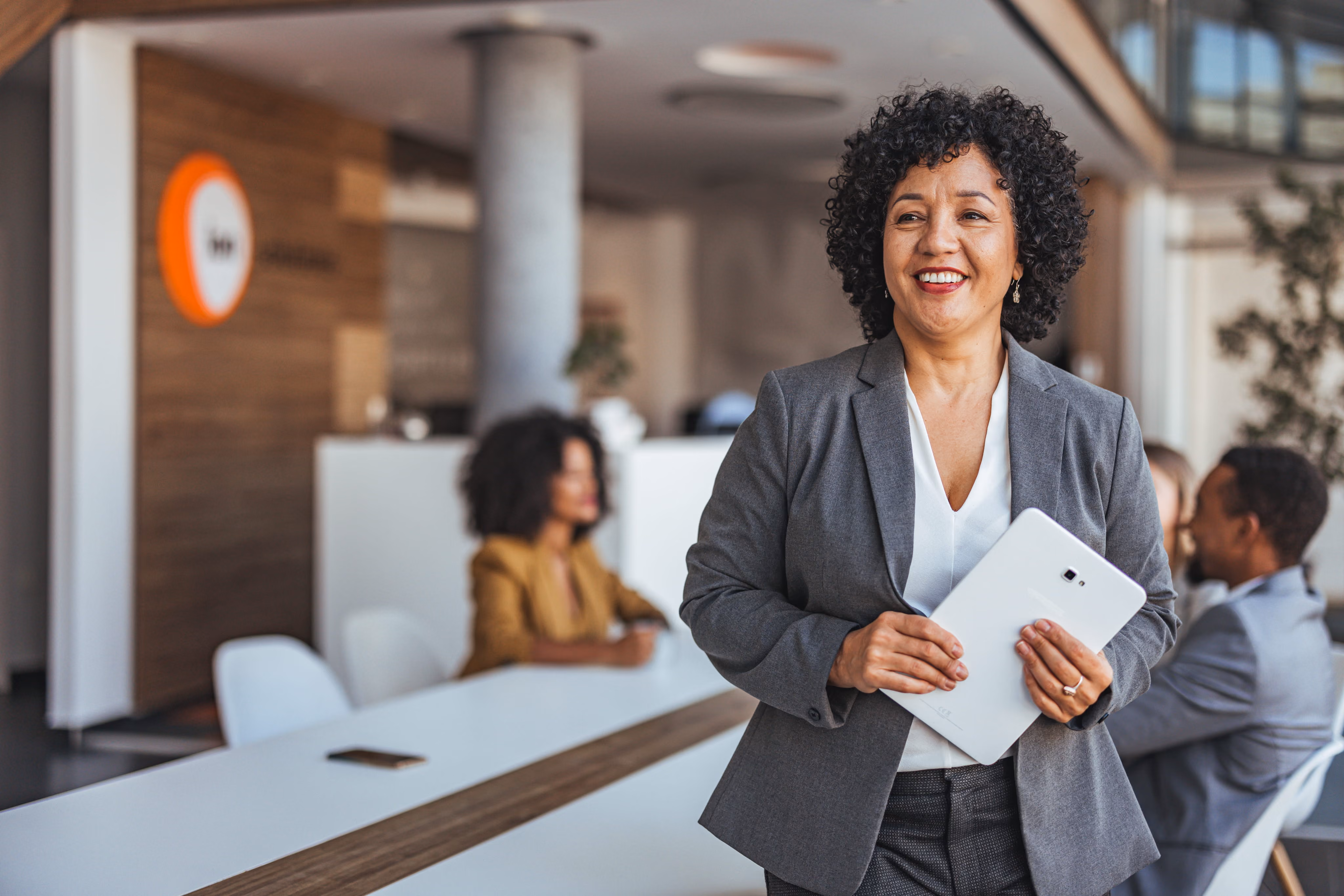 Smiling businesswoman holding a tablet in a modern office with colleagues in the background.