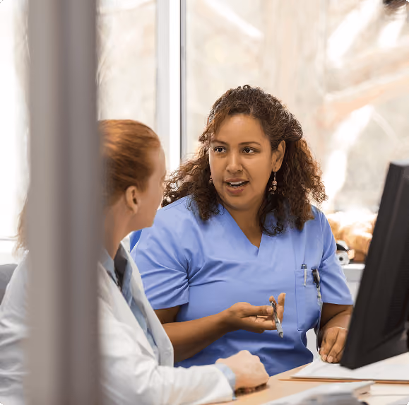 Healthcare professionals in blue and white coats discussing patient info at a computer screen.