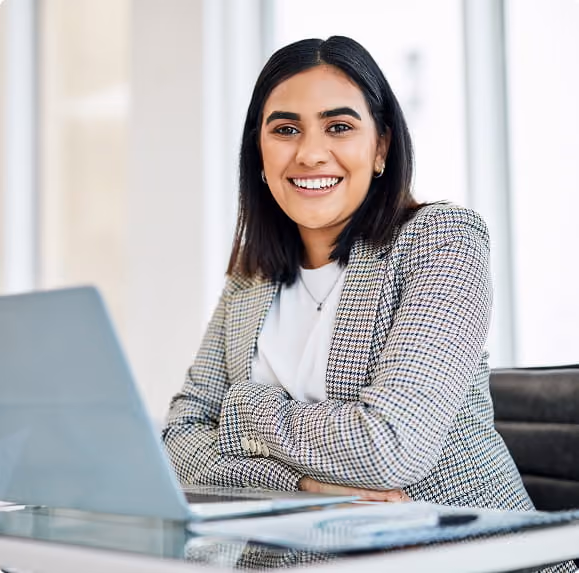 Smiling woman in checkered blazer sitting at desk with laptop in bright office space.