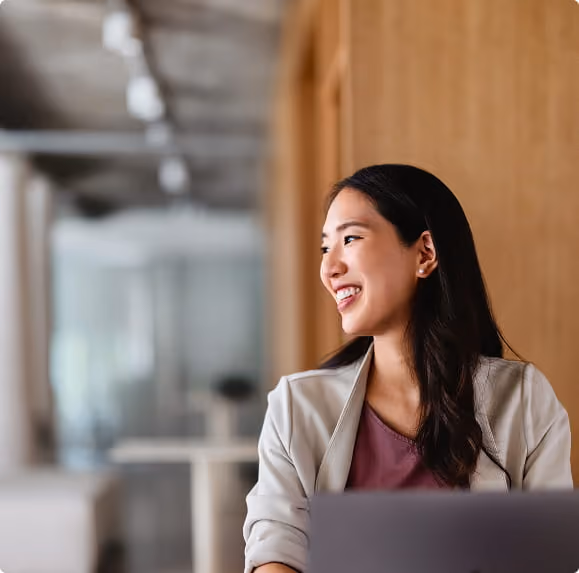Smiling woman sitting and looking to her right with a laptop in front in a modern office.
