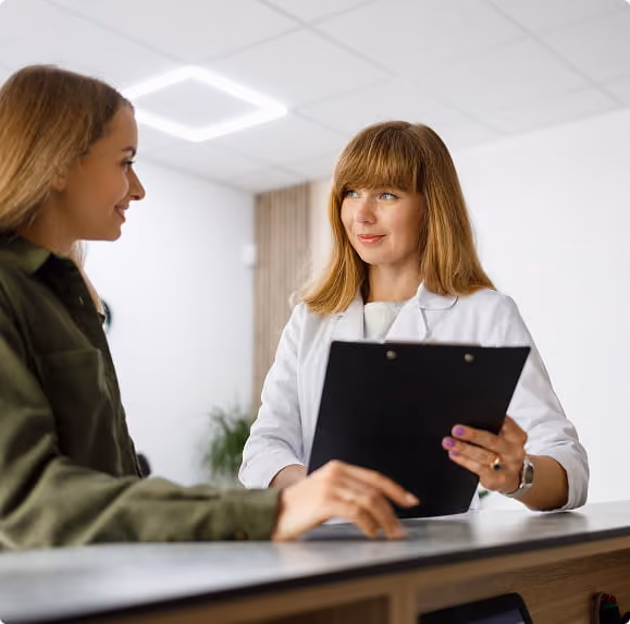 Woman in white coat holding clipboard talking to a smiling woman at reception desk.