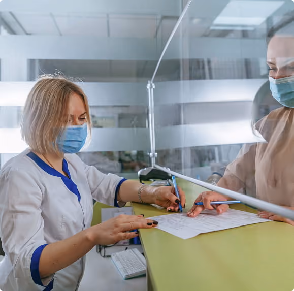 A masked healthcare worker and patient separated by glass window filling out forms with pens.