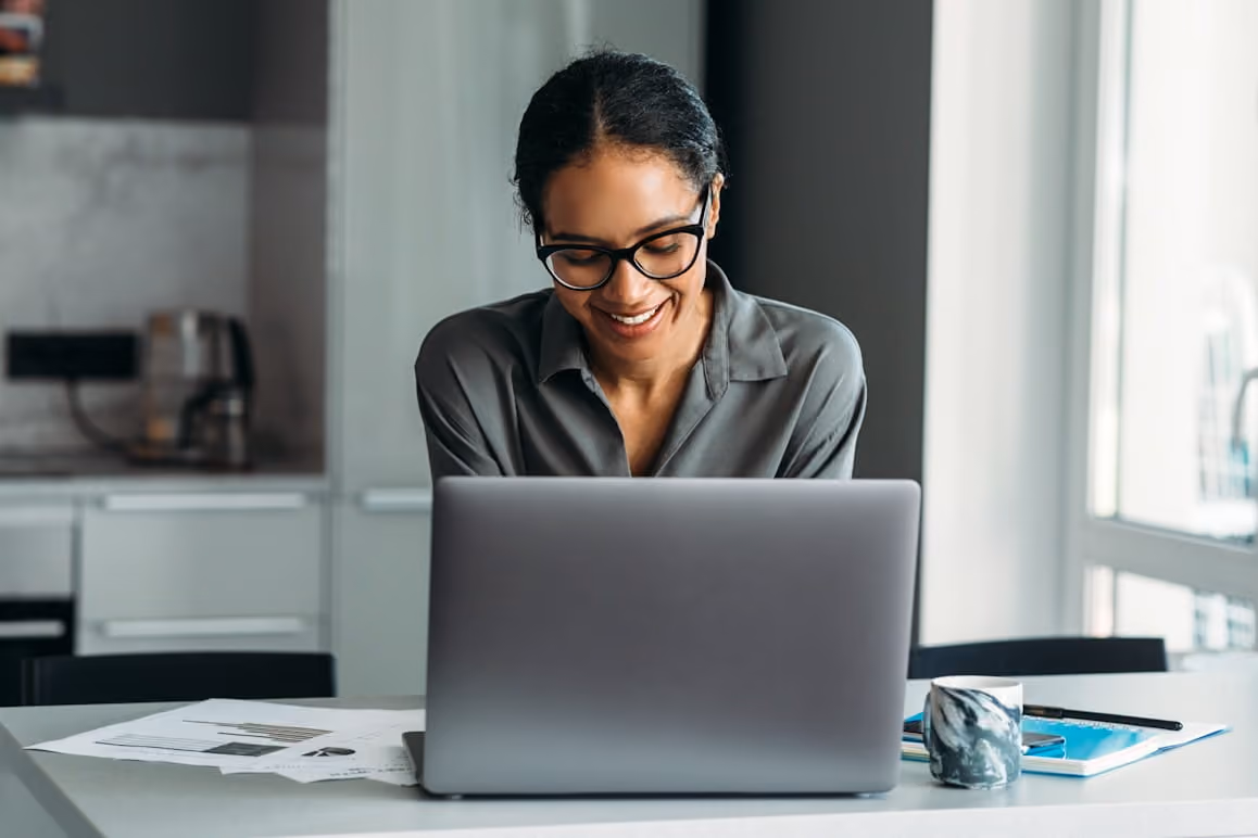 Smiling woman wearing glasses working on a laptop at a table with papers and a mug.