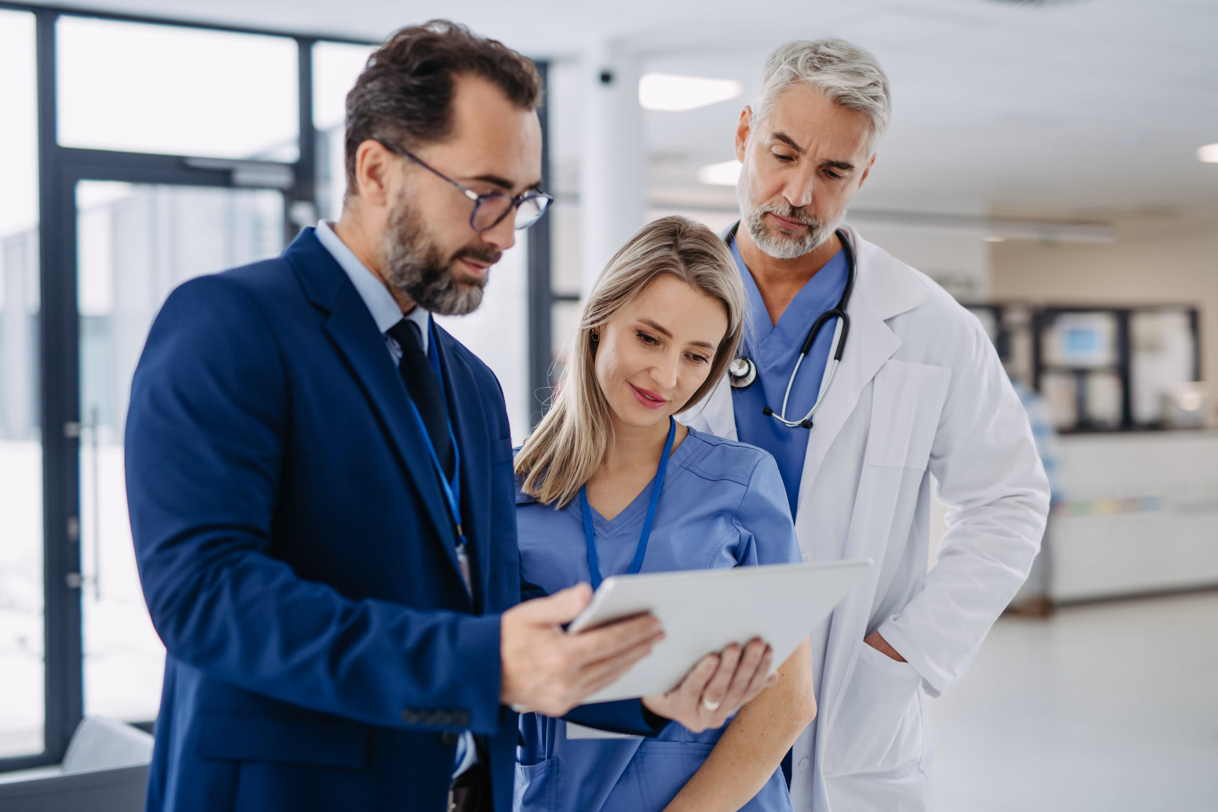 Two medical professionals and a man in a suit reviewing information on a tablet in a hospital hallway.