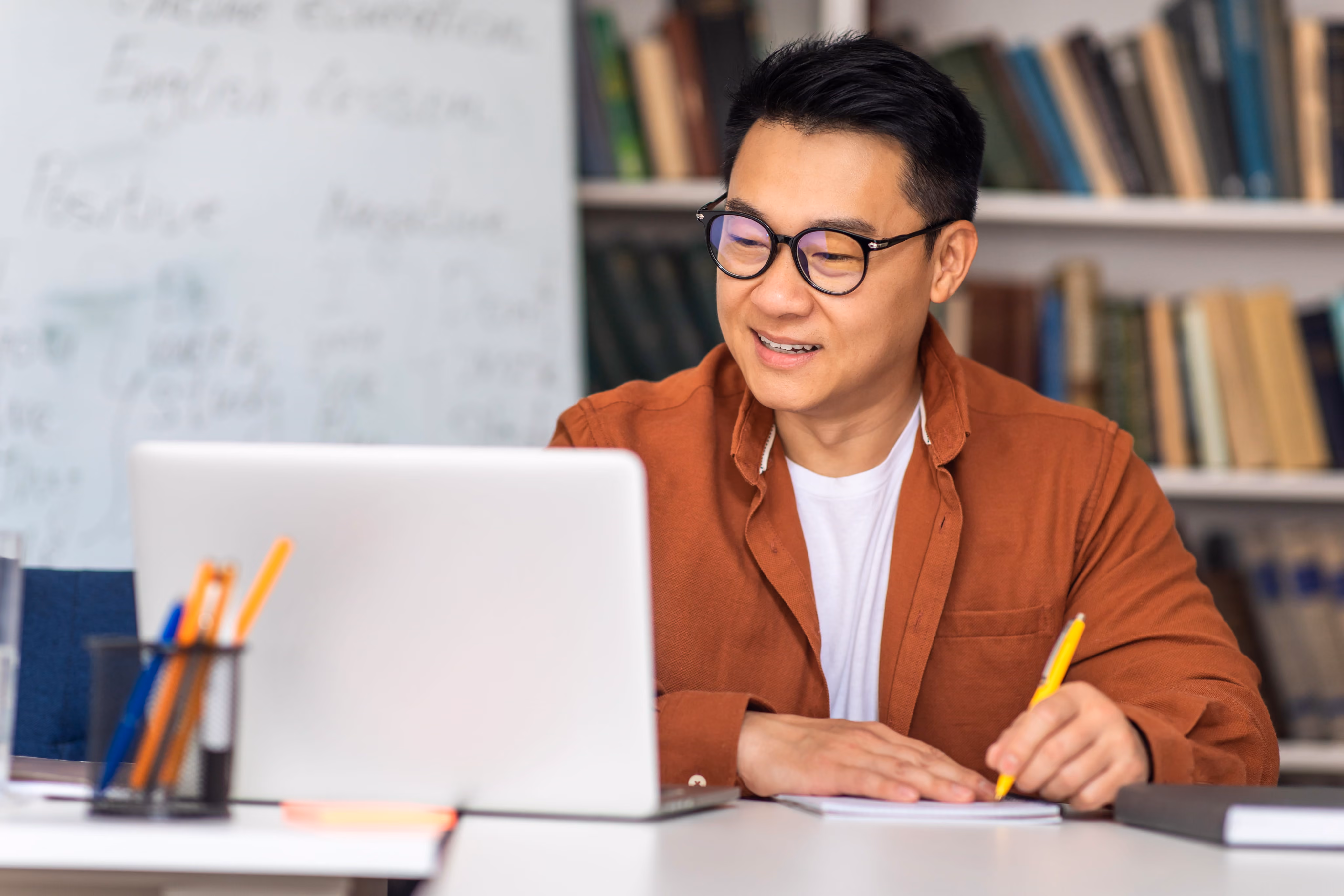 Man wearing glasses and brown shirt smiles while writing notes with pencil near a laptop.