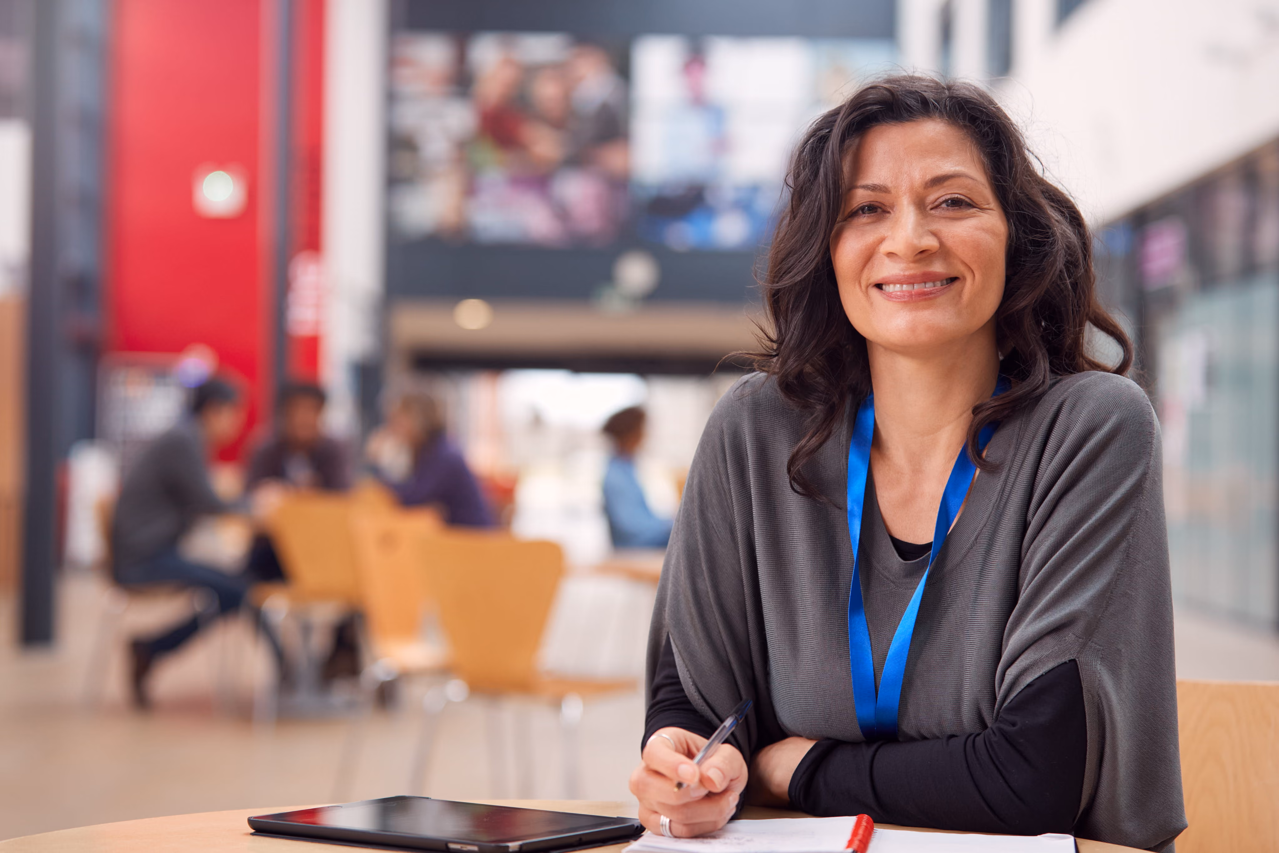 Smiling woman with dark hair in a gray top sitting at a table writing, with tablet and blurred people in background.