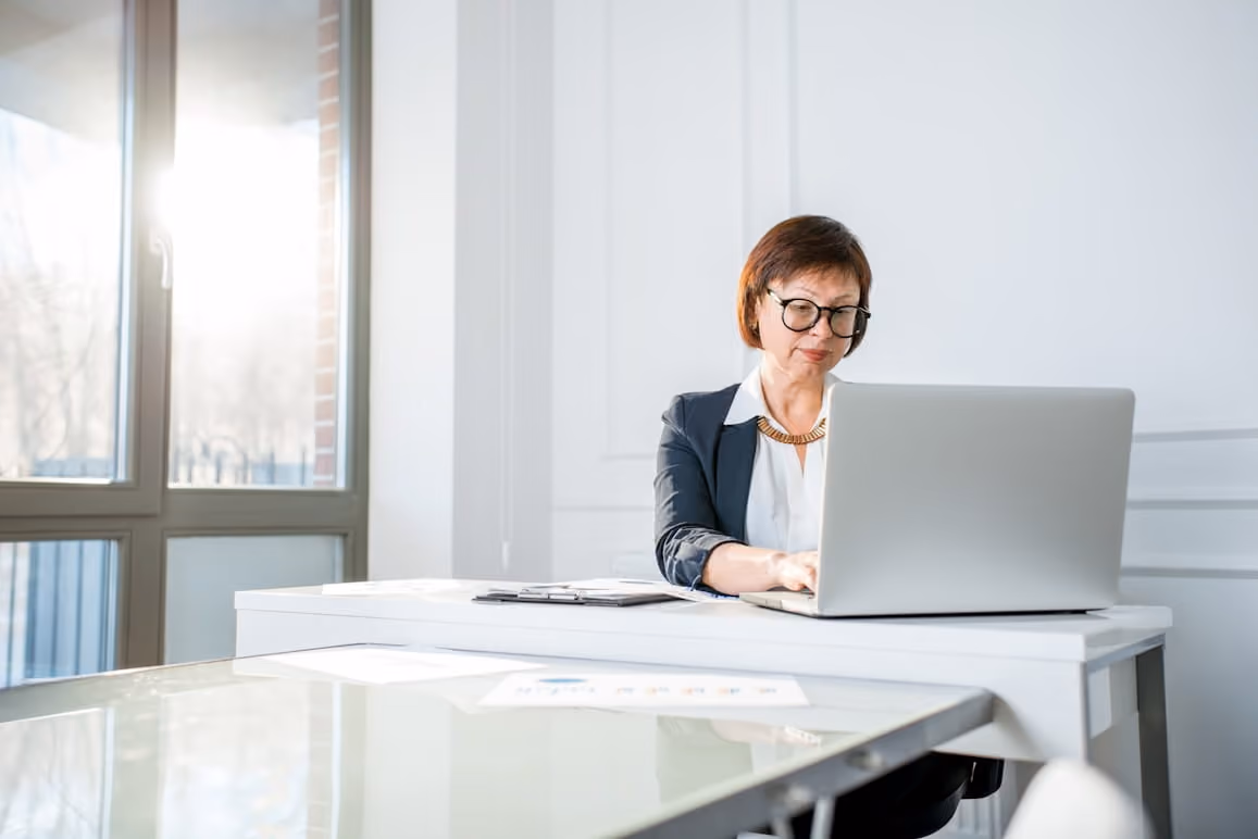 Woman wearing glasses working on a laptop at a white desk in a bright office with large windows.
