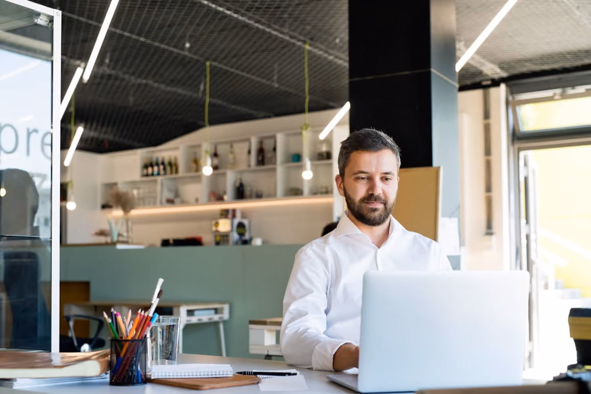 Man with beard working on a laptop in a modern office with shelves and hanging lights.