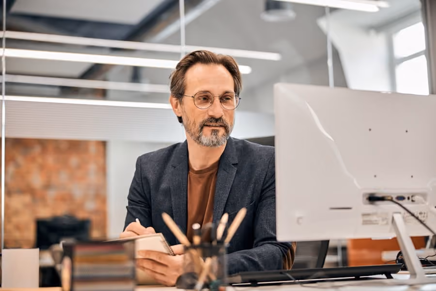 Middle-aged man with glasses writing in notebook while looking at a computer screen in office.