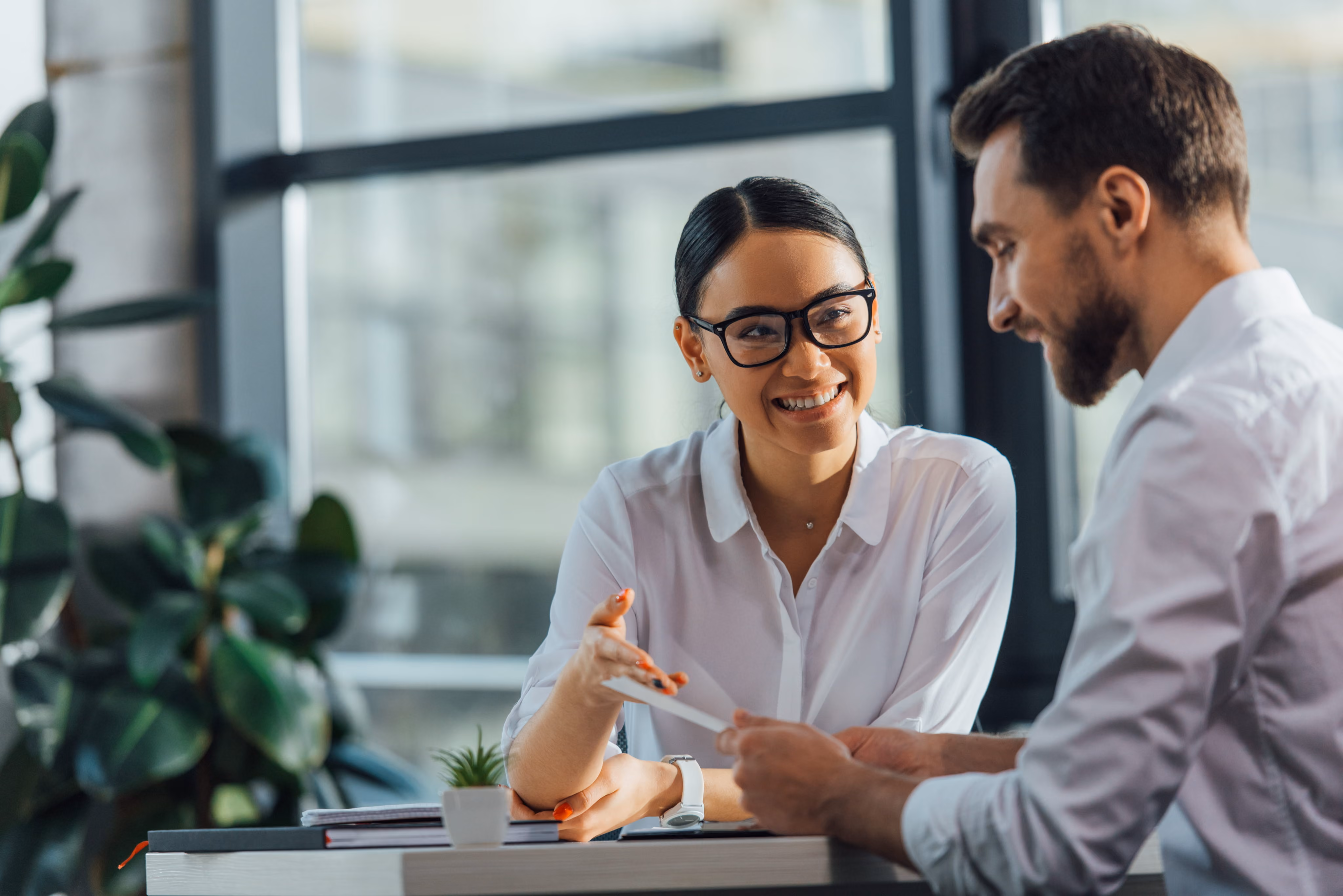 Smiling woman with glasses talks to bearded man at table in a bright modern office.