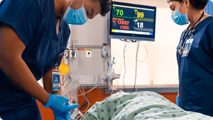 Two nurses in masks attending to a patient lying in a hospital bed with vital signs monitor.