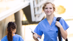 Smiling female nurse in blue scrubs holding clipboard and backpack outdoors near another nurse.