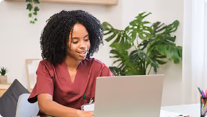 Smiling healthcare worker in scrubs using a laptop at a desk with plants in the background.
