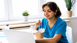 Smiling woman in blue scrubs eating takeout while working on a laptop at a bright desk.