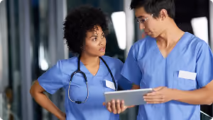 Two medical professionals in blue scrubs discussing information on a digital tablet.