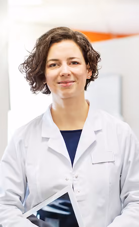 Smiling woman wearing a white lab coat holding a tablet in a bright indoor setting.