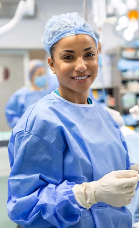 Smiling female surgeon wearing blue scrubs and gloves in a hospital operating room.