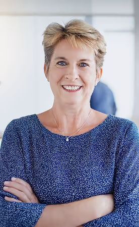 Smiling middle-aged woman with short blonde hair wearing a blue patterned top with arms crossed indoors.